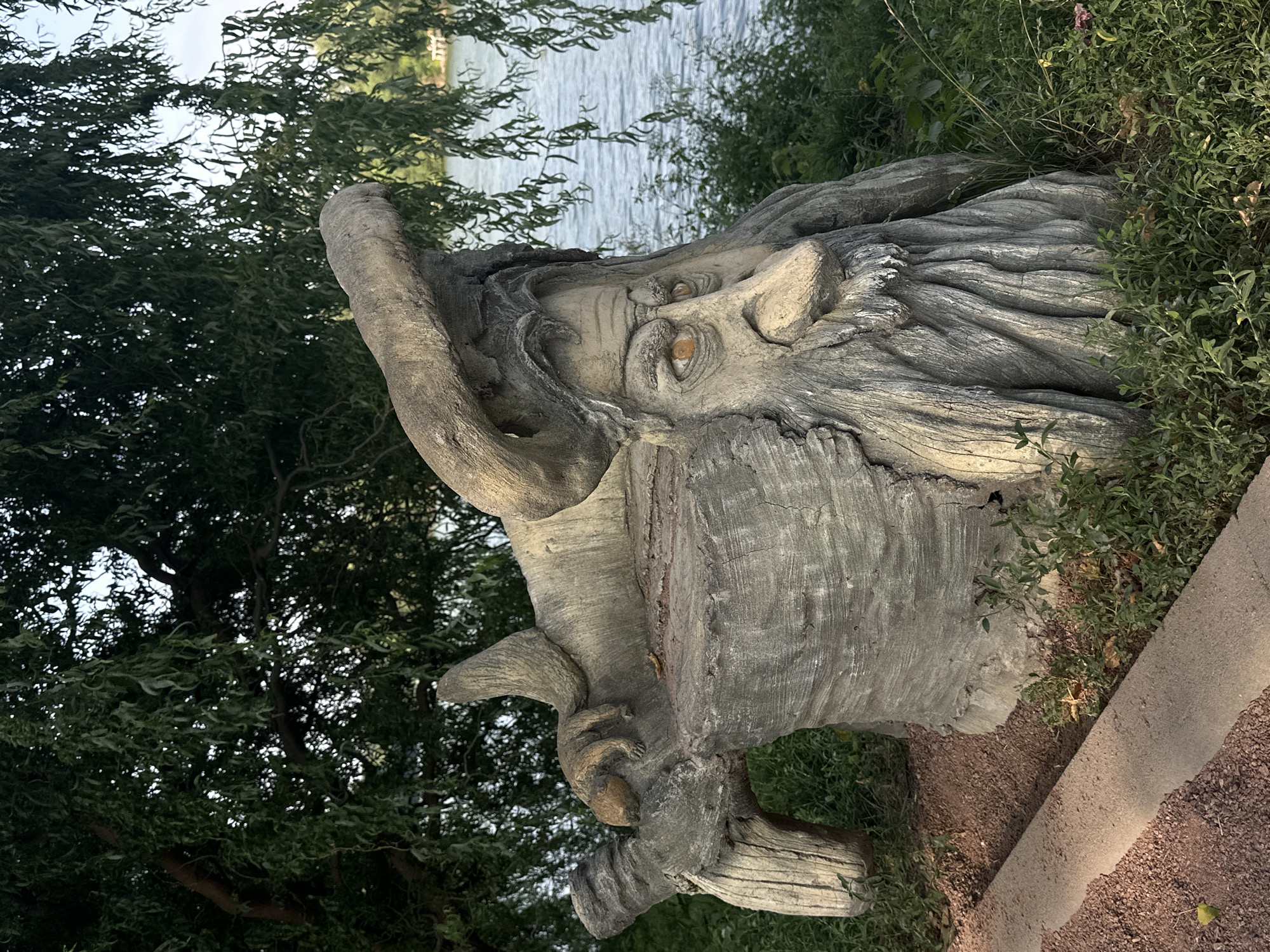 A sculpted stone bench resembling a gnarled tree stump with a face carved into it is situated outdoors near a body of water. The bench has a weathered, grey finish, and the face features a prominent nose, deep-set eyes with orange irises, and a long, flowing beard. A small, sculpted creature, possibly a squirrel or weasel, rests on the armrest of the bench. The background shows lush green trees, foliage, and a calm body of water. A concrete path borders the bench on the left. The lighting suggests it is daytime, likely late afternoon, given the warm tones and shadows. The location is not specified within the city of Unknown, Unknown.