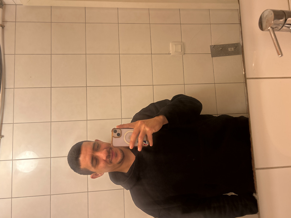 A male individual, appearing to be in his 20s or 30s with short dark hair, is reflected in a mirror, capturing a selfie with a light purple smartphone held in his right hand. He wears a plain black hooded sweatshirt and displays a subtle smile or smirk. The environment is an indoor space, likely a bathroom, characterized by walls covered in white square tiles. Visible in the reflection above the subject is a horizontal silver-colored pipe. To the subject's right, a white electrical light switch is mounted on the tiled wall, and below it, a rectangular metal plate secured by four visible fasteners is affixed. The lower portion of the image reveals the edge of a white ceramic sink and a chrome single-lever faucet. This scene is located in the city of Schiltigheim, France.