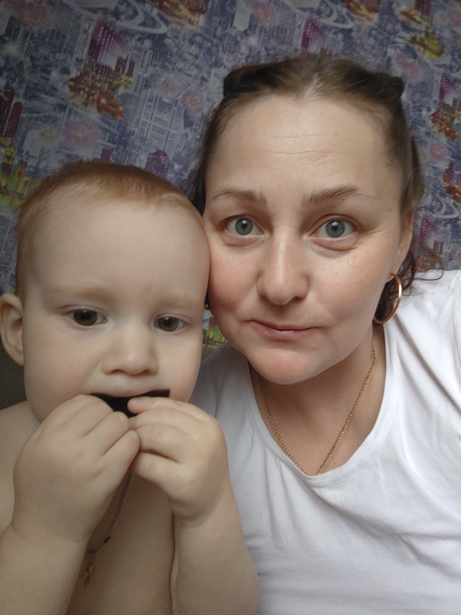 A close-up indoor photograph features an adult woman and a young child. The woman, positioned on the right, has light brown hair pulled back, light blue-green eyes, and a neutral expression as she looks directly at the camera. She is wearing a white t-shirt, a gold chain necklace, and a gold hoop earring is visible on her right ear.
On the left side of the frame, the child, likely a baby or toddler, has light reddish-brown hair and dark brown eyes, looking slightly to the left of the camera. The child's chest is bare, and their hands are brought up to their mouth, holding a dark, rectangular object, possibly a piece of food like chocolate.
The background consists of patterned wallpaper, displaying an abstract design that appears to include stylized buildings or cityscapes in shades of purple, blue, and red. The lighting suggests an indoor setting. No text is visible in the image. The provided context indicates the location is Markovskii, Russia.