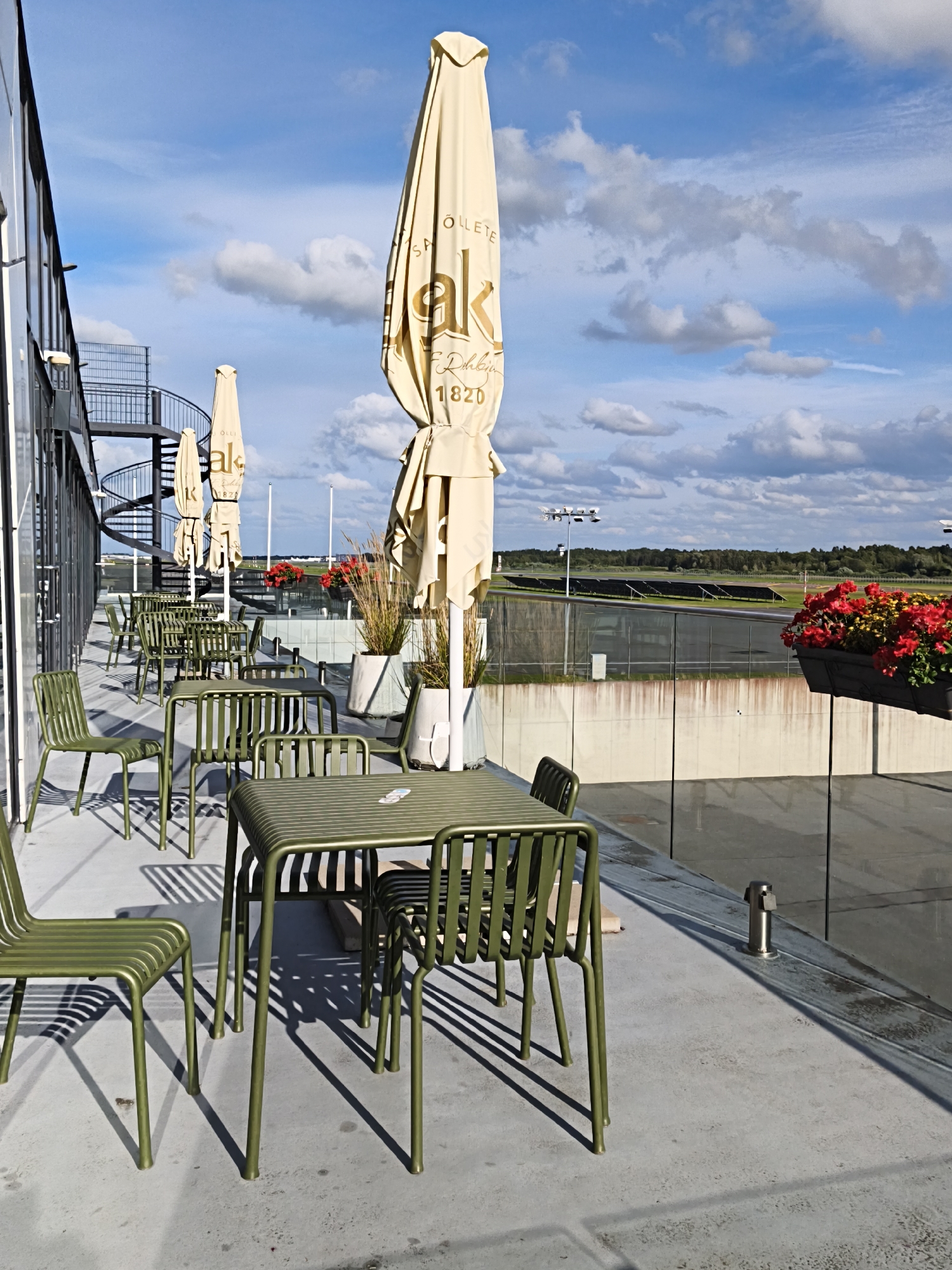 The image shows an outdoor seating area with tables and chairs on a rooftop or balcony. The furniture is a light olive green color. In the foreground, there are several green metal tables and chairs arranged on a concrete floor. Behind the seating area, there are large, closed umbrellas with logos printed on them. A spiral staircase is visible on the left side of the building. In the background, there is a view of what appears to be a sports track or field, with trees and a cloudy blue sky. Red and yellow flowers are in planters along the railing. The scene is set in Tallinn, Estonia.