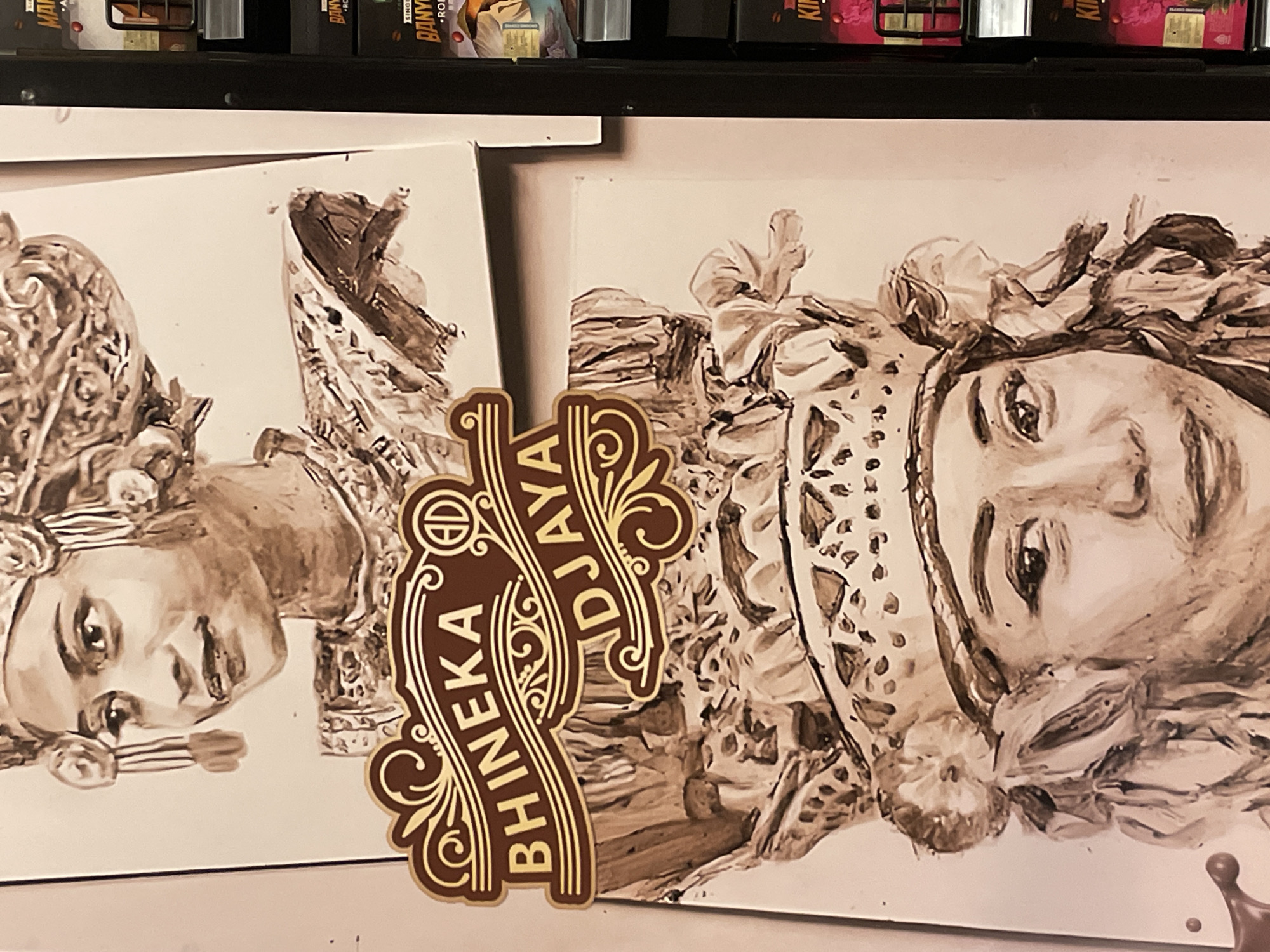 The image displays a static indoor scene featuring two sepia-toned portraits of women, painted with visible brushstrokes on white canvases or panels. The portraits are oriented horizontally within the frame, with the women's heads tilted towards the top edge of the image. The woman in the left portrait has delicate features, gazing upwards with a serene expression, and is adorned with an elaborate, patterned headpiece and an ornate accessory near her mouth. The woman in the right portrait, partially obscured by a foreground element, also looks upwards, wearing a complex head covering, possibly featuring floral or fabric elements.
A prominent brown and gold emblem or logo is centrally positioned in the foreground, overlapping the right portrait. This vertically oriented emblem features intricate scrollwork and the text "BHINEKA DJAYA." A smaller circular logo, containing the letters "BH" and another symbol, is visible at the top of the emblem.
In the blurred background, a dark shelf holds several product boxes. Visible text snippets on these boxes include "BANYU" and "RO" on one, and "KIN" on another, suggesting product names. The setting appears to be an indoor commercial space, such as a shop or cafe, where art is displayed alongside branded products. The overall lighting is warm, enhancing the sepia tones of the artwork and the golden accents of the emblem.