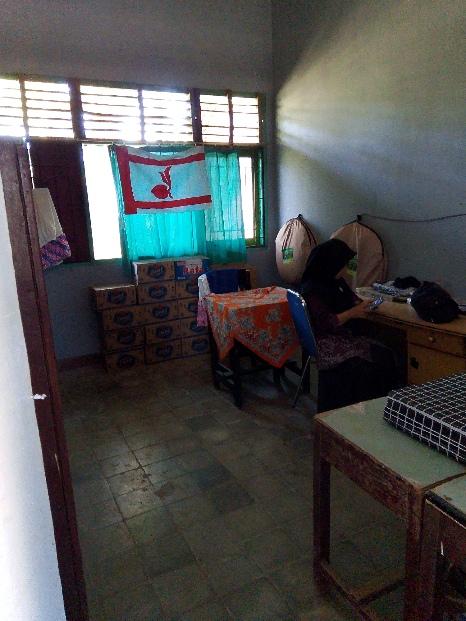 The image depicts an indoor scene, likely a room in a residential or communal setting. In the foreground, a portion of a wooden table with a green surface and a checkered black and white object is visible. To the left, a doorway with a dark brown frame and a white wall is present, with a sliver of white and patterned fabric hanging just inside the frame.
The mid-ground of the image reveals several stacked cardboard boxes, some bearing the text "Deal" and another label that appears to be "Rafa." Adjacent to these boxes is a table draped with a vibrant orange and floral patterned cloth. A person wearing a black hijab and a patterned garment is seated at this table, looking down at a mobile phone. Behind this person, a wooden desk with drawers is visible, holding various items including a black cap and some dark fabric. Two large, light-colored circular objects, possibly bags or cushions, are hanging on the wall behind the desk.
The background features a window with horizontal wooden slats, through which sunlight streams, creating dramatic light rays on the wall. Teal-colored curtains are drawn across part of the window. The floor is tiled with square, gray tiles, some of which appear to have stains or debris. The walls are painted a muted gray-blue color.
The overall impression is one of a somewhat cluttered but functional space. The presence of stacked boxes and the desk setup suggests it could be a storage area, a study space, or a room where goods are being prepared or organized. The light filtering through the window adds a dynamic element to the scene. While the specific location within Kuripan Utara, Indonesia, cannot be definitively determined from the visual information alone, the context implies an Indonesian setting based on the presence of a hijab.