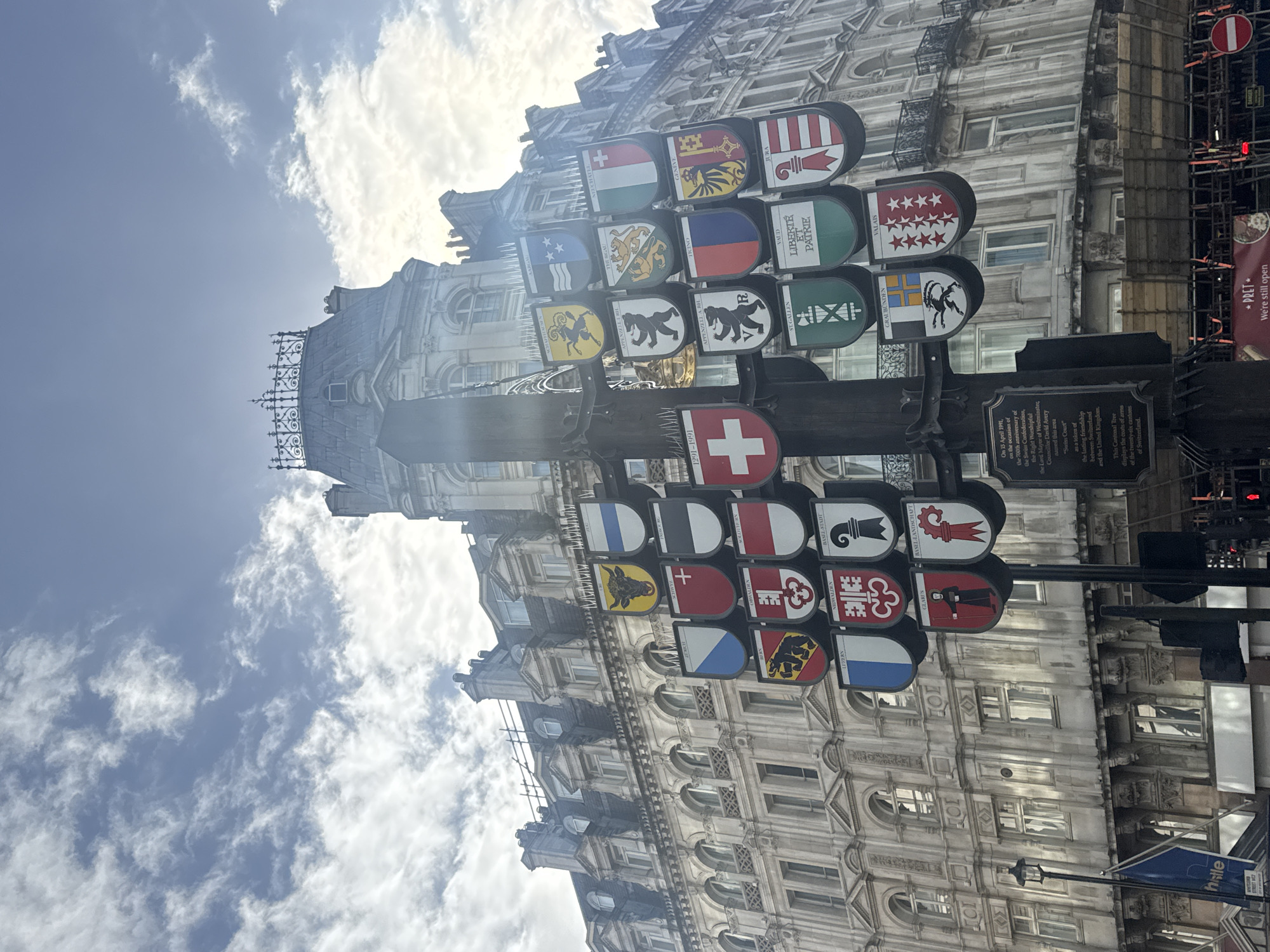 The image shows a Cantonal Tree in Swiss Court, located in the city of London, United Kingdom. The tree is a pole decorated with the coats of arms of the twenty-six cantons of Switzerland. In the background are buildings with ornate facades, partially obscured by the tree. The sky is visible, with a mix of blue and white clouds. A nearby sign indicates that the location is called "Swiss Court" in honor of the friendship between Switzerland and the United Kingdom. There are some other signs including "PRET" which indicates that there is a Pret a Manger shop close by.
