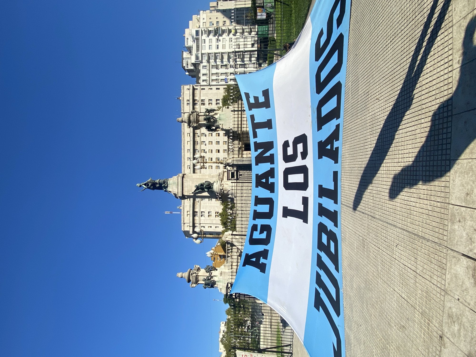 The media file captures an urban outdoor scene, likely a public plaza in Buenos Aires, Argentina, under a clear blue sky. The image is oriented with the sky on the left and the ground on the right, indicating a 90-degree clockwise rotation from a standard landscape view.
Dominating the right side of the frame is a large banner, styled like the Argentine flag with its light blue, white, and light blue horizontal stripes. The banner is hanging vertically, slightly angled, extending from the upper right toward the lower right. Bold black capital letters spell out "AGUANTE LOS JUBILADOS". When the image is viewed correctly (rotated counter-clockwise), "AGUANTE" is on the bottom light blue stripe, "LOS" on the white stripe, and "JUBILADOS" on the top light blue stripe, with the text reading upwards along the banner. This phrase, which translates to "Go retirees!" or "Support for retirees!", strongly suggests a public demonstration, protest, or expression of solidarity for pensioners.
In the background, on the left side of the image, stands an elaborate stone monument featuring multiple tiers, sculptures, and statues, including what appears to be an equestrian figure atop a column. This significant landmark, typical of Buenos Aires architecture, is partially obscured by the banner. Behind the monument and extending further into the background are several multi-story urban buildings with numerous windows, characteristic of a city environment. A metal fence separates the immediate foreground from the background elements.
The paved ground in the bottom right corner features a grid-like pattern and is traversed by long, distinct shadows, suggesting bright sunlight during either early morning or late afternoon. The overall impression is one of a clear, sunny day with a vivid blue sky. The visible text and architectural style provide strong cues to the location being a public square in Buenos Aires.