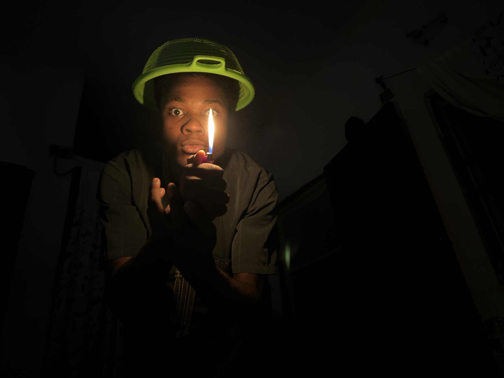 A young person wearing a green colander on their head and a dark t-shirt holds a lit lighter in front of their face, illuminating their eyes and mouth. The background is dark, with hints of furniture or draped fabric visible. The person's expression is one of surprise or shock. The scene appears to be indoors, likely in a residential setting, and the lighting suggests it's nighttime or the room is dark. The location of Ihiagwa, Nigeria is not discernible from the image.