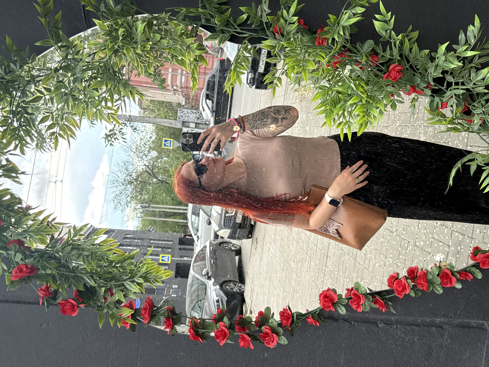 A woman with red hair is reflected in a round mirror decorated with greenery and red roses. She wears sunglasses, a beige top, a black skirt, and a watch. She holds a smartphone in her right hand, seemingly taking a picture. Her left hand rests on a brown purse. Both arms have tattoos. The mirror reflects a street scene in Rostov-on-Don, Russia, including cars, buildings, a pedestrian crossing sign, and a cloudy sky.