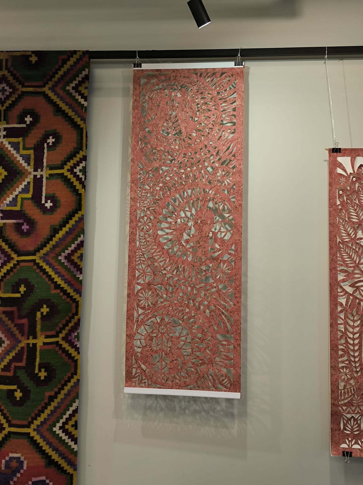 The image shows an indoor scene with a patterned textile hanging on the left and two coral-colored, intricately cut paper artworks hanging vertically on a light grey wall. The patterned textile features geometric shapes in shades of green, yellow, purple, and orange. The paper artwork in the center is a long rectangle with floral and geometric cutouts. It is suspended by clips and hangs from a black track mounted on the ceiling. Another similar paper artwork is partially visible on the right. A black track light is positioned on the ceiling above the central artwork. The location is potentially an art gallery or museum, although the exact location in Vinnytsia, Ukraine cannot be confirmed from the image alone. There are no people visible in the scene.