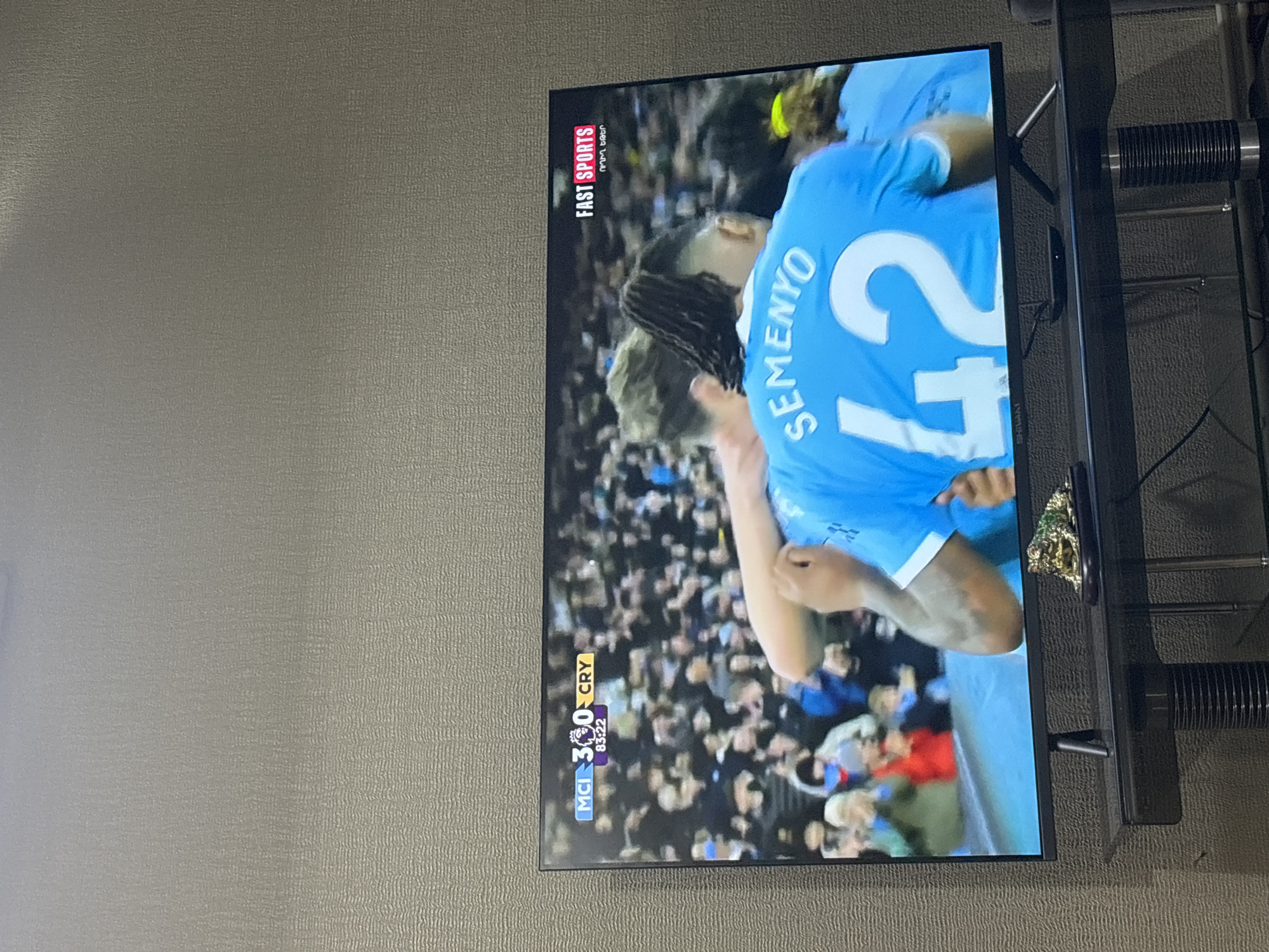 The image shows a television screen displaying a football match between Manchester City (MCI) and Crystal Palace (CRY). The score is 3-0 in favor of Manchester City, with the match time at 83:22. The player on the screen has the name "SEMENYO" and the number "42" on his jersey. The screen also displays the "FAST SPORTS" logo. The television is mounted on a black stand with a glass shelf. The background wall is a light gray color. The location is in an unspecified area, Yerevan, Armenia.