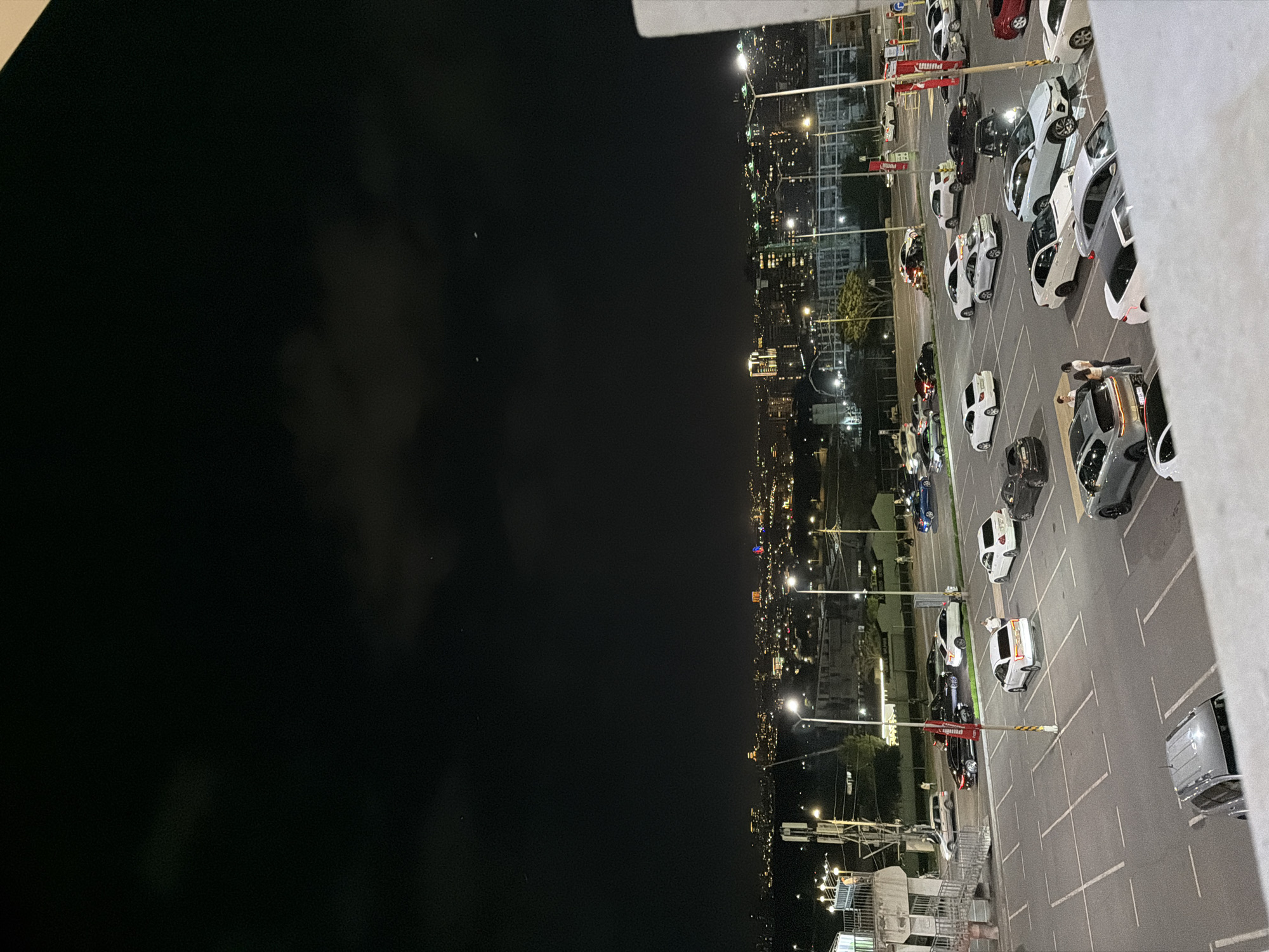 The image depicts a nighttime view of a parking lot and the city of Yerevan, Armenia. The parking lot is filled with various cars, mostly white and black. Streetlights illuminate the lot and the adjacent cityscape, which is visible in the distance. The city lights create a blurred effect due to the distance. The sky above the city is dark.