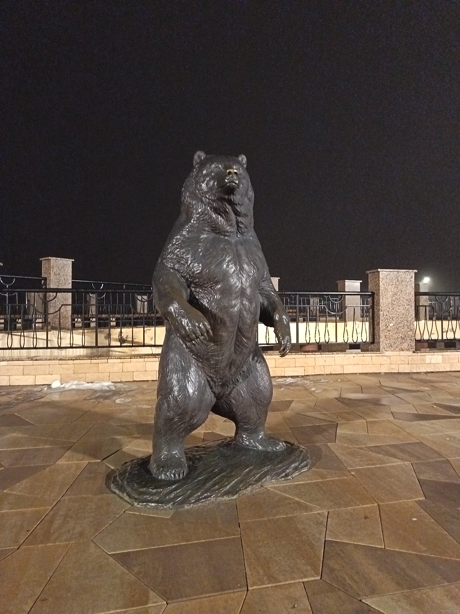 A bronze statue of a bear standing upright on a pedestal is centered in the image, positioned on a paved square in the city of Nizhnevartovsk, Russia. The bear is the primary subject, facing forward with detailed fur texture. In the background, a black wrought iron fence with decorative elements runs along a low beige wall, potentially a promenade or walkway. Behind the fence, stone pillars with lights on top are spaced out. The ground is paved with large, geometric stones. The sky is dark, suggesting it's nighttime, and the scene is artificially lit. Small patches of snow or ice are visible near the fence.