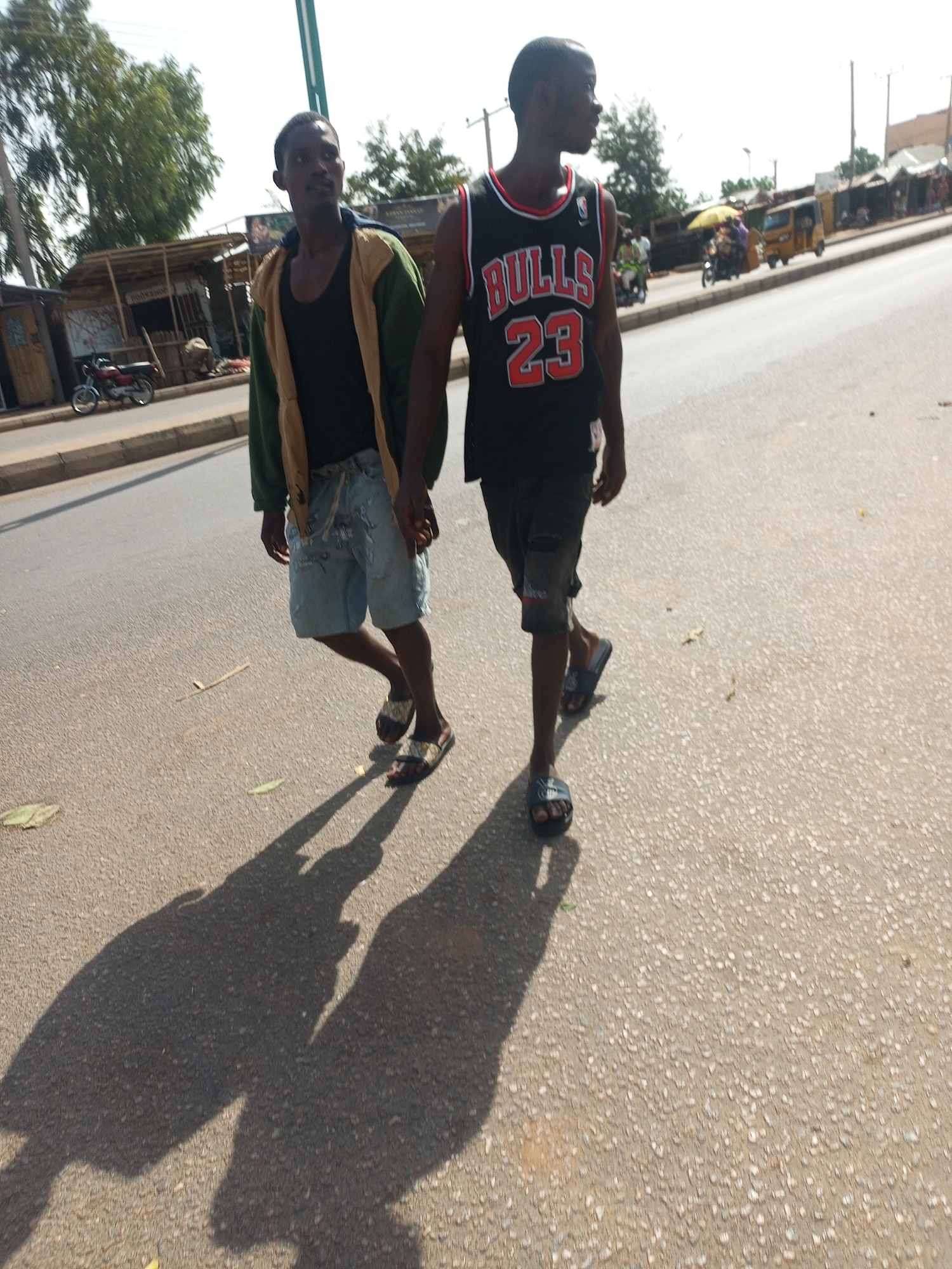 Two young men are depicted walking along the side of a paved road in Tempure, Nigeria, under a bright, sunny sky. The long shadows cast by the subjects suggest it is late morning or afternoon.
The man on the right wears a black basketball jersey featuring "BULLS 23" in red and white lettering, paired with dark shorts and black slide sandals. He is looking forward or slightly to his right. The man on the left wears a black tank top under an open green and tan jacket, light blue distressed denim shorts, and patterned slide sandals. He appears to be looking forward. His right hand is close to the other man's left hand, implying they might be walking hand-in-hand.
The scene showcases a typical street environment. Along the left side of the road, there are several rustic stalls or small shops with wooden structures. A motorcycle is parked near one of these structures. Further down the road, various vehicles, including a yellow auto-rickshaw (commonly known as Keke Napep in Nigeria) and other motorcycles, indicate ongoing traffic. Green trees line the background, and the sky is clear and bright. The road surface is asphalt, separated by a curb from the dirt area where the shops are situated.
Visible text includes "BULLS 23" on the jersey of the man on the right. A faint sign reading "BOOKSHOP" can also be seen on one of the background stalls. The image captures an ordinary moment of daily life on a lively street.
