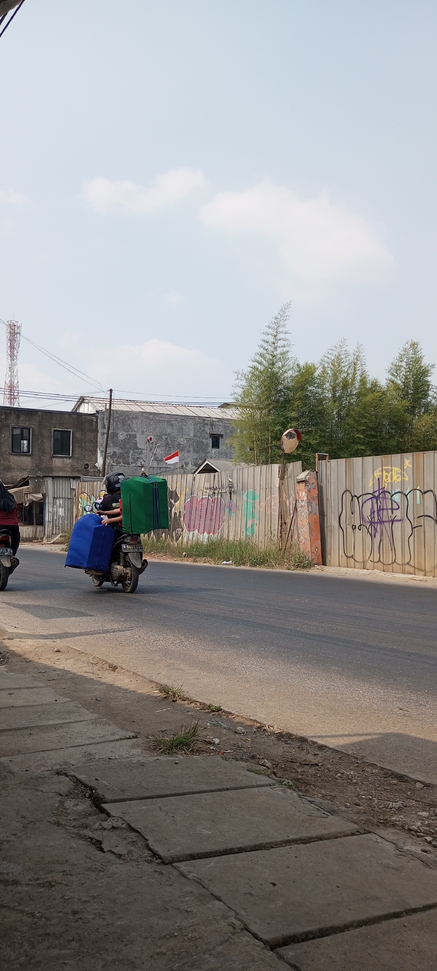 A person wearing a black helmet rides a scooter with two large boxes strapped to the back, one blue and one green, on a paved road in Setu, Indonesia. The scooter is moving away from the camera, to the right. The road is bordered by a concrete sidewalk on the left and a metal fence covered in graffiti on the right. Behind the fence, there are trees and buildings, including a cell tower. The sky is clear and blue with some scattered clouds. The scene appears to be an urban or suburban environment, likely during the daytime, under bright sunlight.