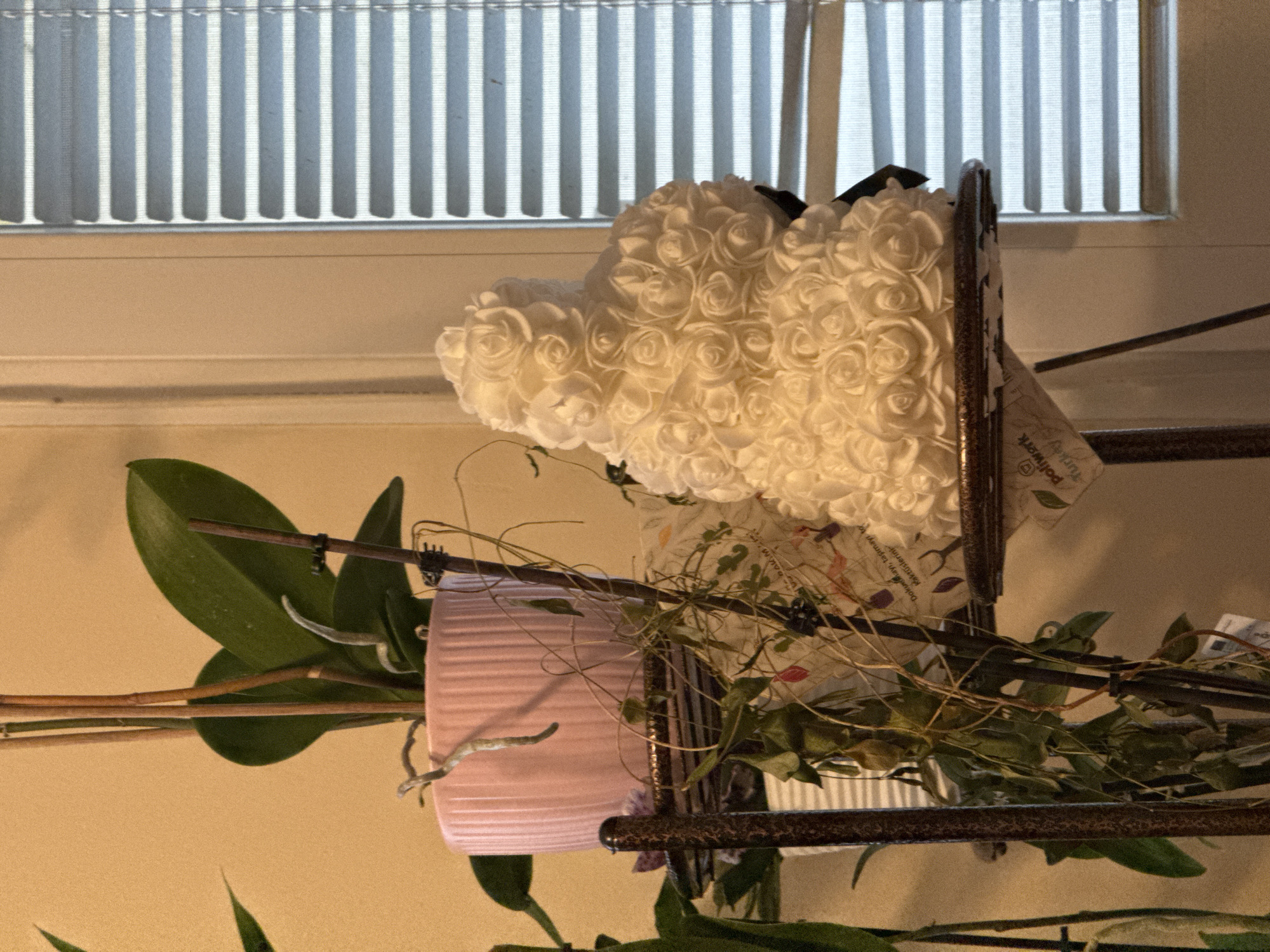The image shows an indoor scene with a shelf holding a pink pot with orchid leaves, a white heart-shaped arrangement of roses, and some other greenery. The shelf appears to be made of metal rods. In the background is a window covered with vertical blinds. There is no evidence to suggest that the location is Sofia, Bulgaria.