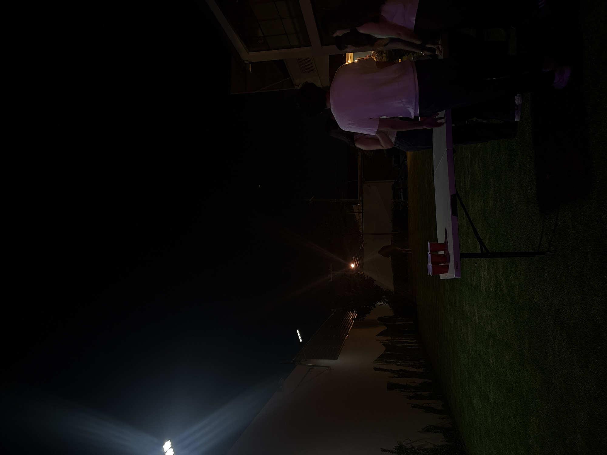 At night in San Bernardino Tlaxcalancingo, Mexico, a group of individuals appears to be engaged in a social gathering outdoors on a lawn. A white folding table is set up, with red cups, suggesting a party or game of beer pong. Two people are standing at the table, one wearing a white shirt, while others are nearby. The background includes a building or structure, and streetlights illuminate the scene.