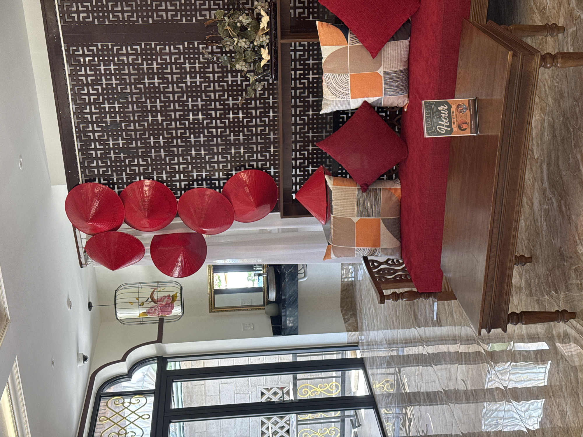 The image captures a scene inside a building in Đà Nẵng, Vietnam. The room is decorated with traditional Vietnamese elements, including a row of red conical hats arranged along the top of a wall. A decorative wooden screen with a patterned design spans part of the wall behind the hats. The scene includes a seating area with a red cushion sofa and decorative pillows in red and geometric patterns. A wooden table is placed in front of the sofa, with a book on top. Outside, visible through a large arched window, there's a view of an external area with a decorative metalwork fence. The lighting suggests a bright, daytime scene. A mirror on the wall in the background reflects some of the interior. The room has a tiled floor. No people are visible in the image.