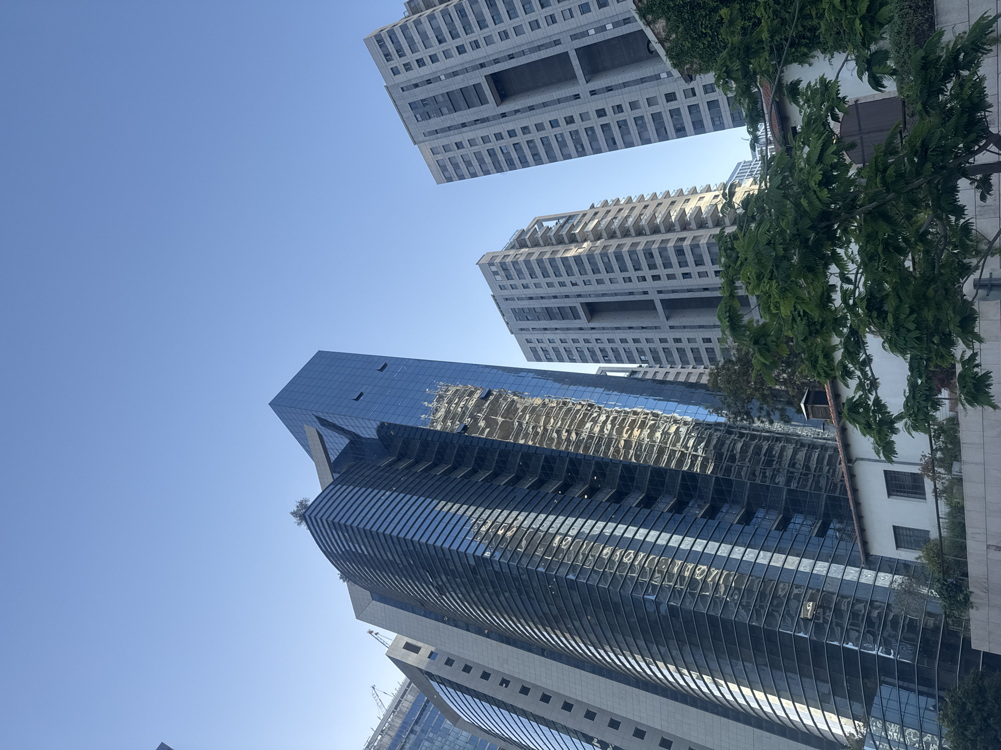 The image presents a modern urban landscape in Tel Aviv, Israel, captured from a low-angle perspective looking upwards against a clear, bright blue sky.
The scene is dominated by several contemporary high-rise buildings. In the foreground and extending towards the mid-ground, a prominent dark-colored skyscraper features a distinctive curved, ribbed glass facade. This building's reflective surface mirrors the surrounding environment, showing inverted images of other buildings and the sky. To its right, another glass-clad building with a more angular design is partially visible.
In the upper portion of the frame, two striking light-colored buildings with unique architectural styles are present. These appear to be multi-story structures, possibly residential or commercial, characterized by a grid of windows, geometric patterns, and large, rectangular cut-outs within their design. Some green foliage, possibly ivy, clings to the side of one of these buildings.
On the right side of the image, vibrant green trees with dense leaves provide a natural contrast to the urban architecture. These trees frame the buildings, suggesting well-maintained streetscapes or parks within the city.
The bright, cloudless sky and clear light indicate a sunny day, likely mid-morning or afternoon. No people or specific activities are discernible in the scene, which captures a static architectural view. There is no visible text in the image.