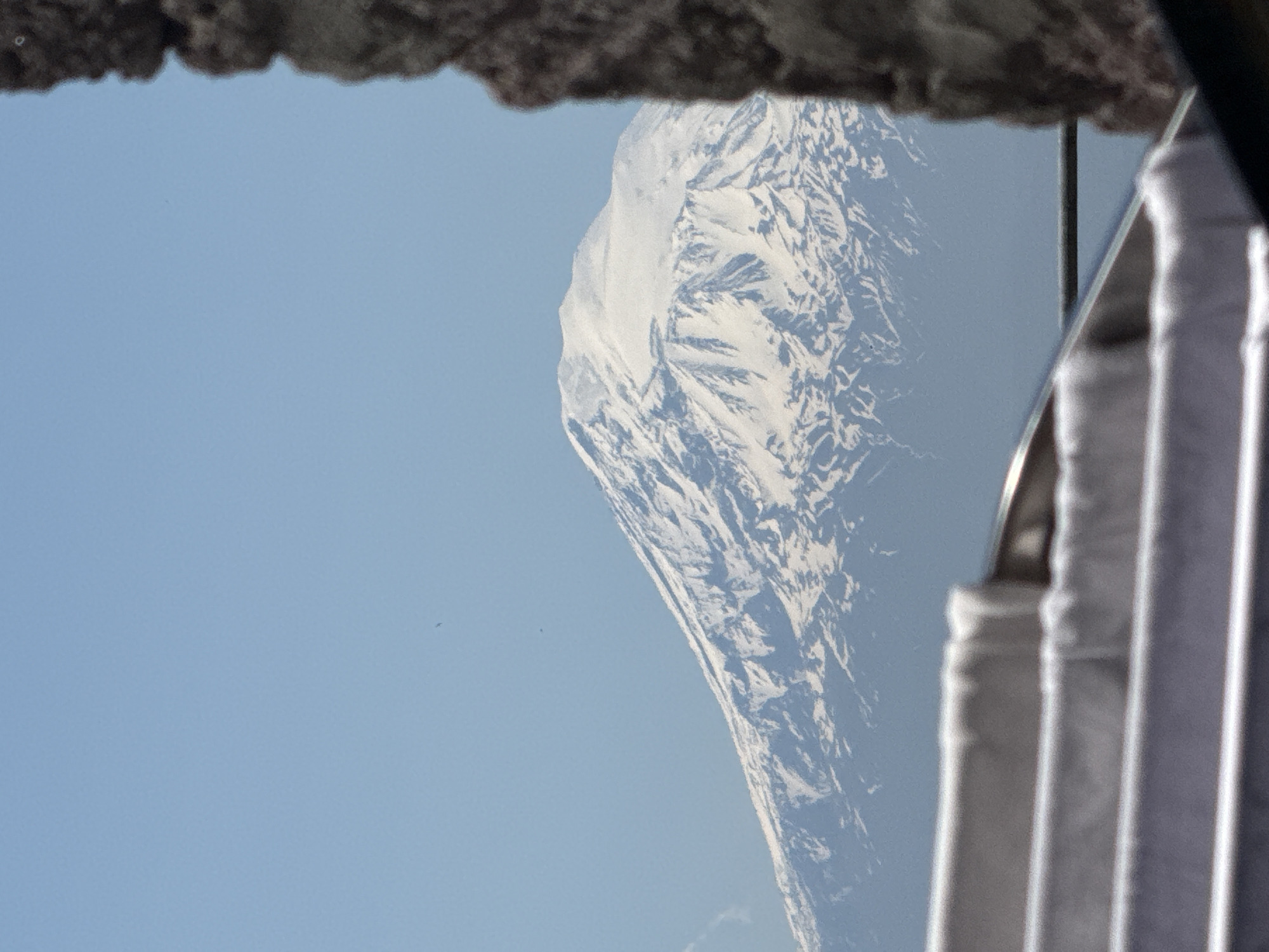 The image, oriented correctly, depicts a vast, snow-capped mountain under a clear, pale blue sky. The mountain, rugged and imposing, occupies the central and right portions of the frame, its extensive slopes and peaks covered in white snow and ice, revealing intricate textures and shadowed crevices. The sky above and to the left of the mountain is uniformly blue and free of clouds, suggesting bright, clear weather.
In the foreground, at the very top, a dark, textured, and possibly rocky or concrete overhang partially frames the view. On the right side, blurred vertical elements made of metallic or reflective material are visible. These elements resemble a window frame, a railing, or part of a reflective surface, indicating that the mountain is being observed from an enclosed or semi-enclosed structure, such as a building or an observation deck. A small, dark speck, possibly a distant bird or a dust particle on the lens, can be seen against the clear sky in the upper left quadrant.
The scene captures a moment of serene natural beauty and grandeur, likely taken during the daytime due to the bright and consistent illumination. No people or specific events are visible, only the majestic landscape and elements of the viewing environment. There is no discernible text in the image.