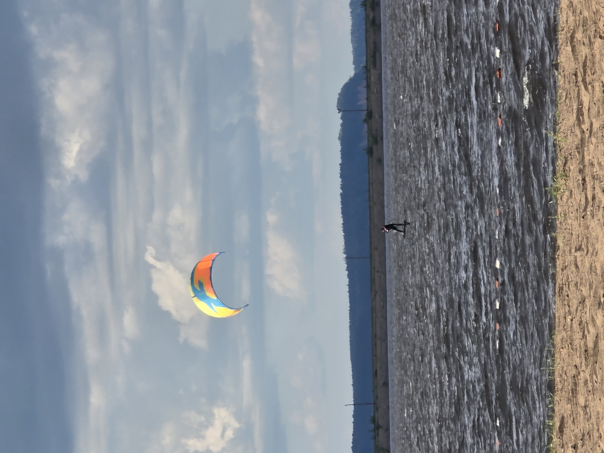 A lone kitesurfer is visible on the dark, choppy waters of the Kama River in Chaikovsky, Russia. The kitesurfer is in the middle distance, positioned towards the right side of the frame. Above the water, a brightly colored kite, featuring yellow, orange, and blue sections, arches gracefully against a cloudy sky. In the background, a dense line of dark green trees forms a distant horizon, with a faint power line visible across it. The foreground consists of a sandy beach. A line of buoys, alternating between white and orange, stretches across the water in the middle ground. The overall scene depicts a recreational activity on a large body of water under an overcast sky.