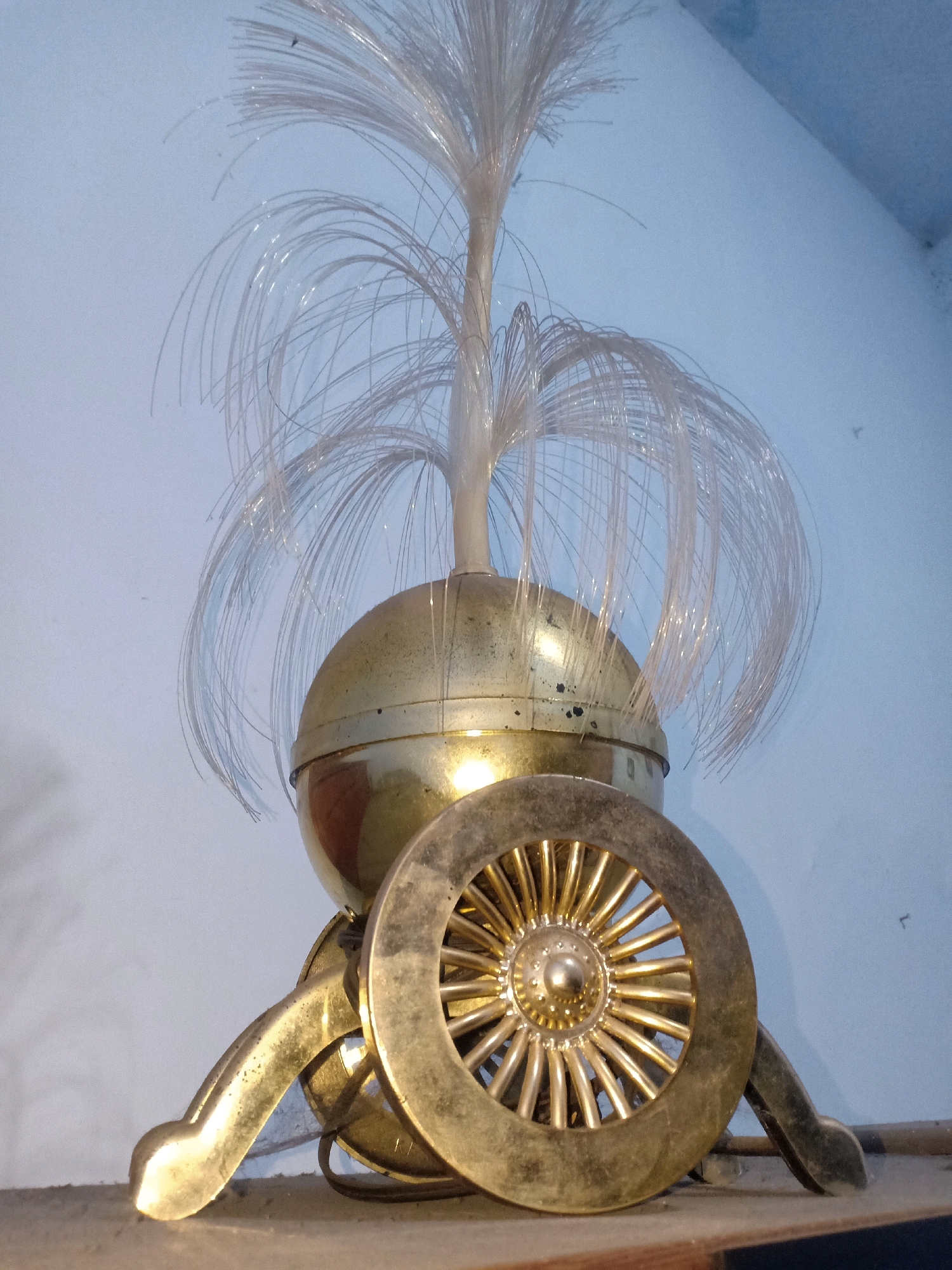 An ornate, brass-colored object with a spherical base topped with a cluster of translucent fiber optic strands that fan out like a plume. The object has a decorative wheel in the foreground with numerous spokes radiating from a central hub. The entire object is positioned on a shelf in front of a light blue wall. No people or any other objects are visible. The scene is indoors. The image quality is fair, with some graininess and slight blurriness. The lighting appears to be artificial. The object is likely a decorative lamp or a part of a larger art installation.