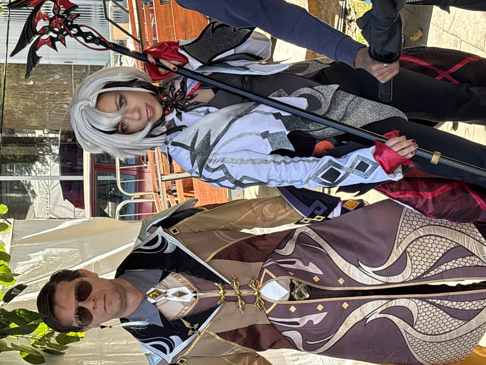 Two cosplayers are posing outdoors, possibly at an event. The person on the left is a man dressed in an ornate brown and gold costume with a high collar and a long, flowing cape. He is wearing aviator sunglasses and a decorative earring. The person on the right is a woman with short, white and black hair, dressed in a white and black costume with red accents. She is holding a staff with a red and black, spiky head. Her costume features a detailed collar and patterned sleeves. Their outfits appear to be inspired by characters from a video game or anime. They are standing in front of a white tent structure and what looks like outdoor seating, possibly in a park or public space. The background includes some greenery and sunlight, suggesting it is daytime. There is another person on the far right, partially visible, wearing a blue sweater and holding a black object, possibly part of another costume. The location is described as being in the city of São Paulo, Brazil.