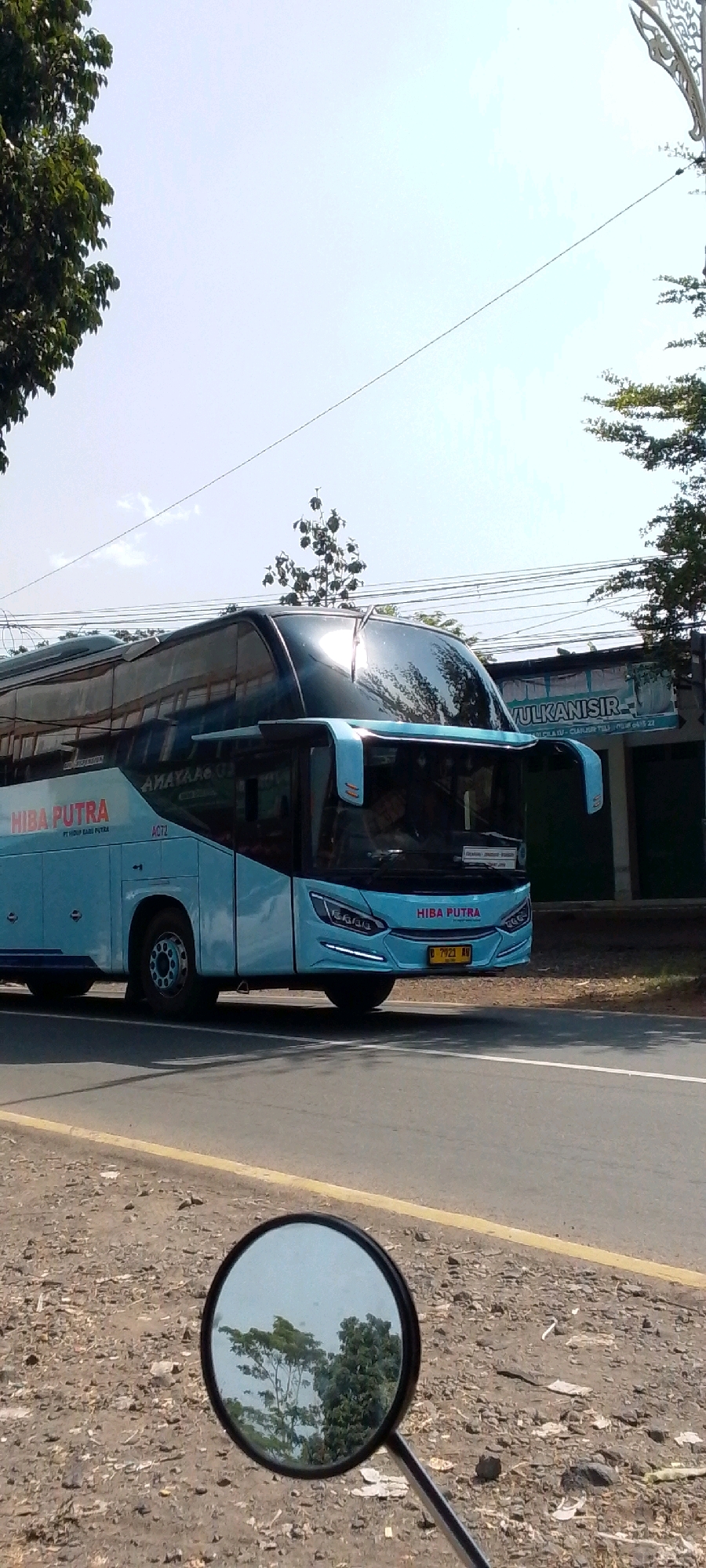 This is a vertical, eye-level, medium shot of a light blue intercity bus on a paved road. The bus, identified by the "HIBA PUTRA" logo on its side and front, is traveling to the right. The front of the bus features modern headlights and a yellow license plate with the characters "D 7921 AV". The bus has tinted windows, and reflections of trees and the sky are visible on its surface.
To the right of the bus, a building with a sign that reads "VULKANISIR" is partially visible. The road has a yellow line marking the shoulder. The ground beside the road is covered in gravel and debris. In the foreground, a motorcycle's round side mirror is visible, reflecting trees and the sky. The overall environment suggests an outdoor setting in Cilaku, Indonesia, likely on a sunny day, given the clear sky and the sharp reflections.