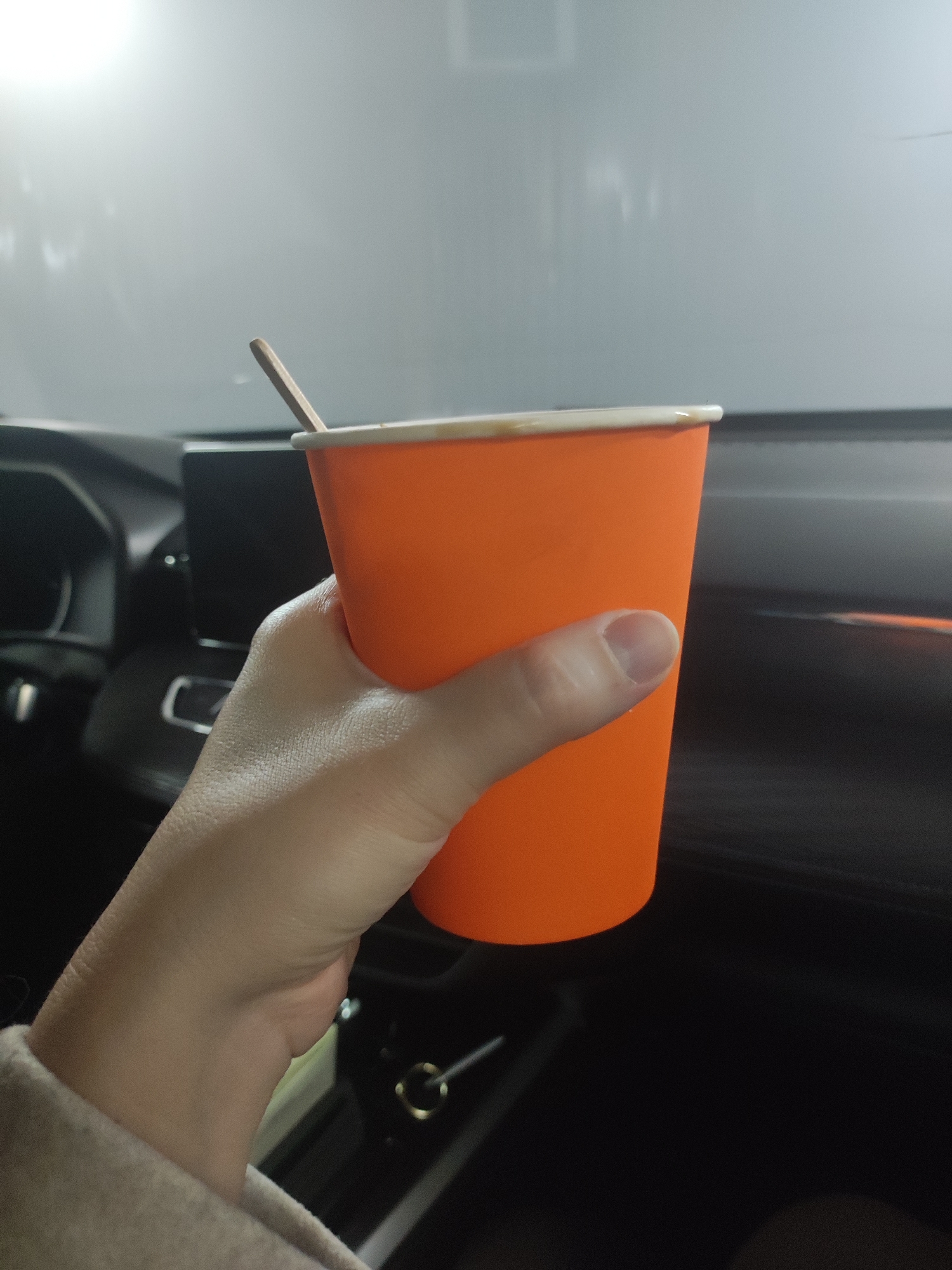 A hand is holding an orange paper cup with a wooden stirrer inside. The cup is likely filled with a beverage. The scene is inside a vehicle, evidenced by the dashboard and steering wheel visible in the background. The location is unknown and cannot be reliably determined as Votkinsk, Russia.