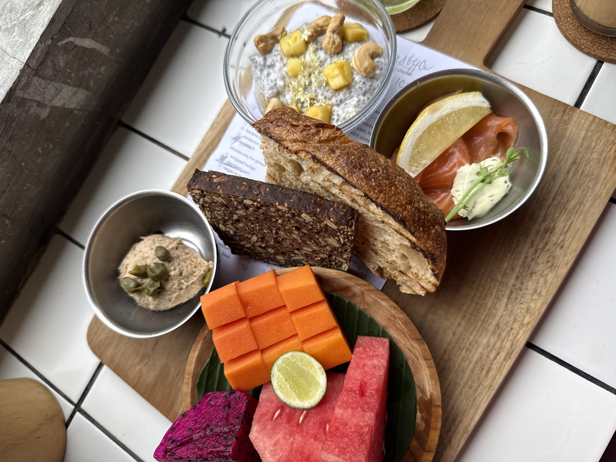 A high-angle, eye-level close-up shows a wooden serving board laden with a variety of food items, set against a background of white tiled surfaces, potentially in North Kuta, Indonesia. The main focus includes a clear bowl containing a white, seedy substance, topped with diced mango and cashews. Slices of brown and lighter bread lean against this bowl. Adjacent is a small metal bowl with smoked salmon, a lemon wedge, and what seems to be a dollop of cream cheese garnished with greens. Another metal bowl holds a beige-colored spread with green capers.
Below this arrangement is a circular wooden dish filled with fruit: cubed papaya, wedges of watermelon, a slice of dragon fruit, and a lime. A green banana leaf serves as a base for these fruits. The corner of what seems to be a printed menu or ingredient list is visible, and a cylindrical container sits in the background, possibly holding a drink. The tiles suggest an indoor setting, likely in a café or restaurant.
