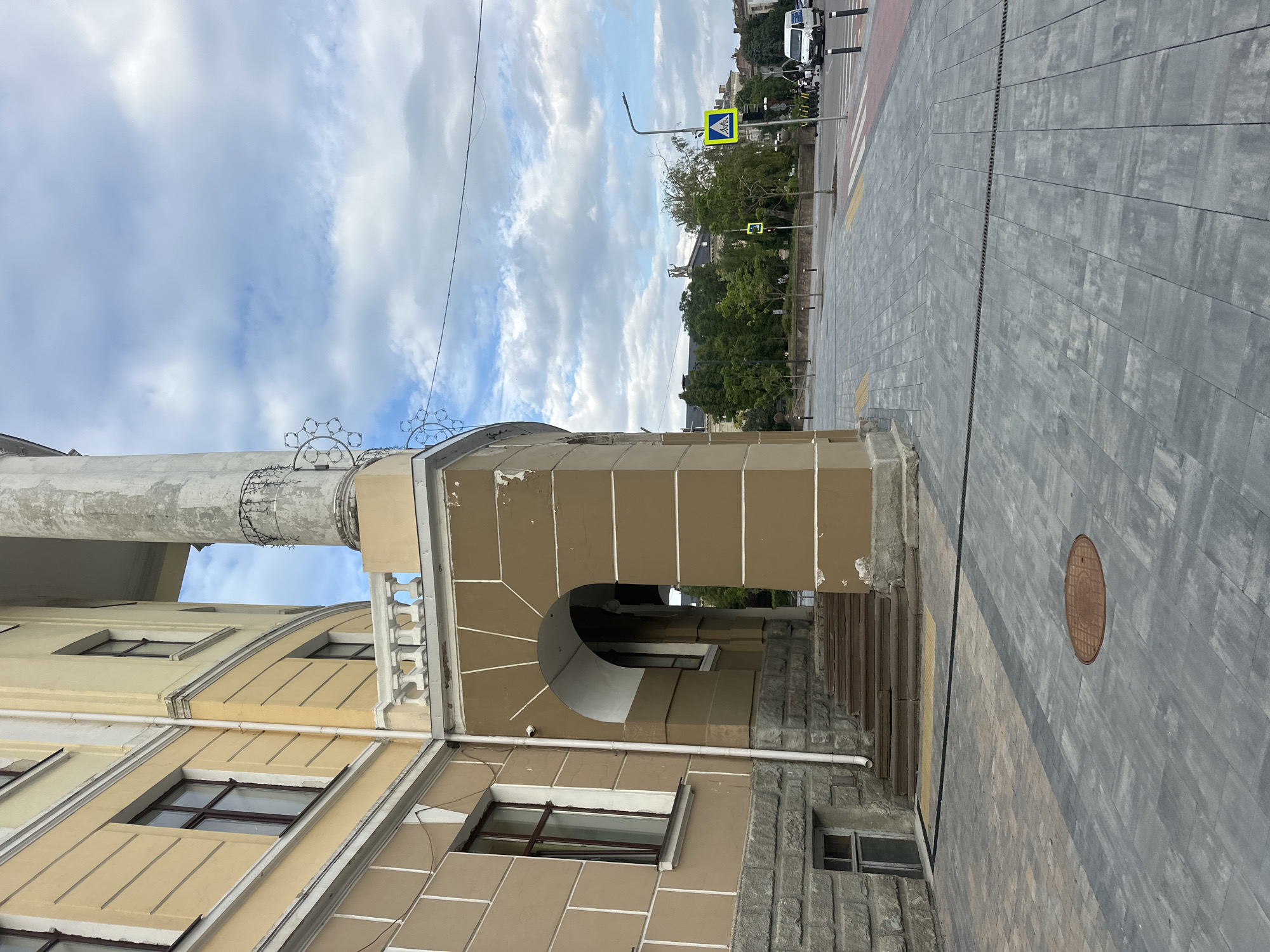 This is a medium-angle, eye-level, outdoor, vertical shot of a building facade and sidewalk in Ternopil, Ukraine. The building, painted a light yellow and brown, features classical architectural elements, including a large column and arched entrance. Steps lead up to the arched entrance, with a small, barred window visible at the base of the building on the left. The sidewalk is paved with grey and tan interlocking stones, with a manhole cover visible in the foreground. Across the street, a pedestrian crossing marked with red and white stripes is visible, as are several yellow scooters, a white van, and a pedestrian crossing sign. Trees and other buildings form the background under a cloudy blue sky. The scene appears to be a public area, likely in a city center or historical district.