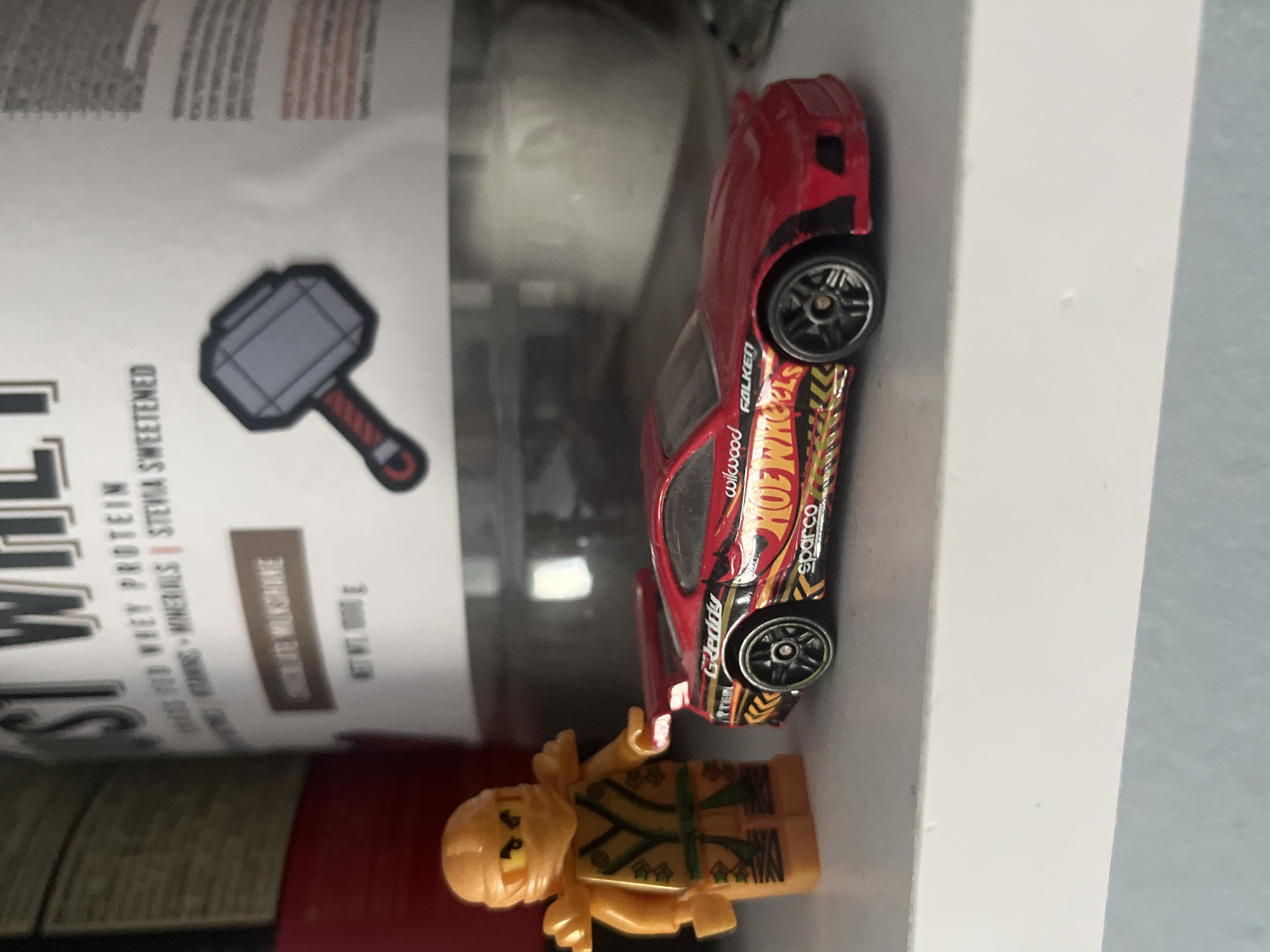 In Pokrovnik, Bulgaria, a still life photograph captures a collection of objects on a white shelf. On the left, a Lego minifigure of a ninja, predominantly orange and gold with green accents, stands beside a red toy car. The toy car, a Hot Wheels model, features intricate racing decals, including "GReddy," "Falken," "Wilwood," and "Sparco." Its design suggests a performance-oriented vehicle, with a prominent rear spoiler and custom wheels.
Behind the toys, a large white tub of "JUST WHEY PROTEIN" is visible, adorned with text in black and red. Notably, a stylized depiction of Thor's hammer, Mjolnir, is present on the tub's label, rendered in grey and red. The background is blurred, hinting at a domestic or storage environment, possibly a home in Pokrovnik. The lighting is moderate, casting soft shadows and highlighting the textures of the subjects. No people are present in the scene, and the arrangement of objects suggests a display or collection.