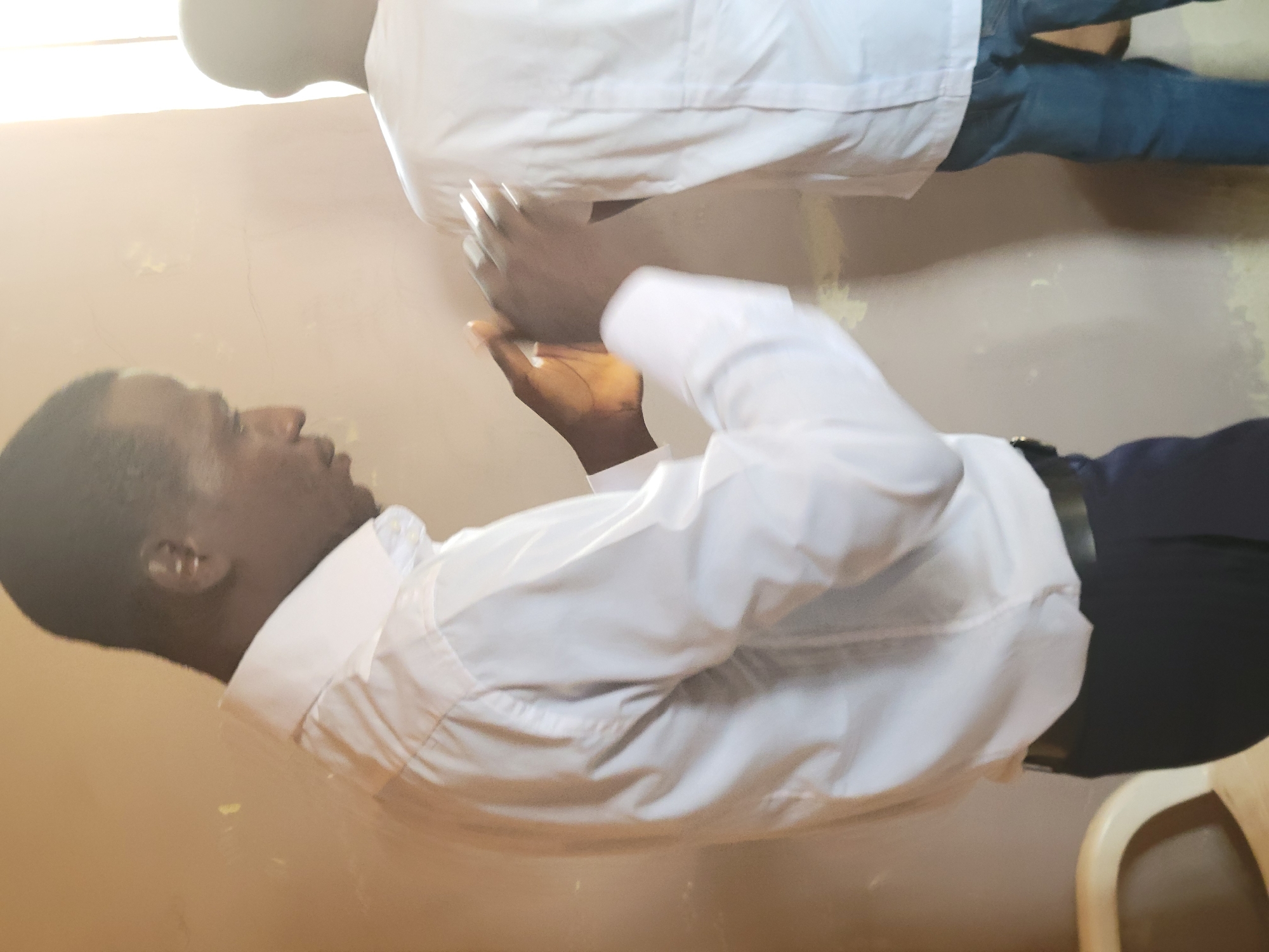 A sideways indoor shot shows a dark-skinned man in a white, long-sleeved shirt, black pants, and a black belt. His head is toward the left side of the frame, and he is looking downward with his hands together. Another person is seen above him, also wearing a white shirt and blue jeans. The setting is simple, with a light-colored wall and what appears to be a chair. The location is reported as Madalla, Nigeria.