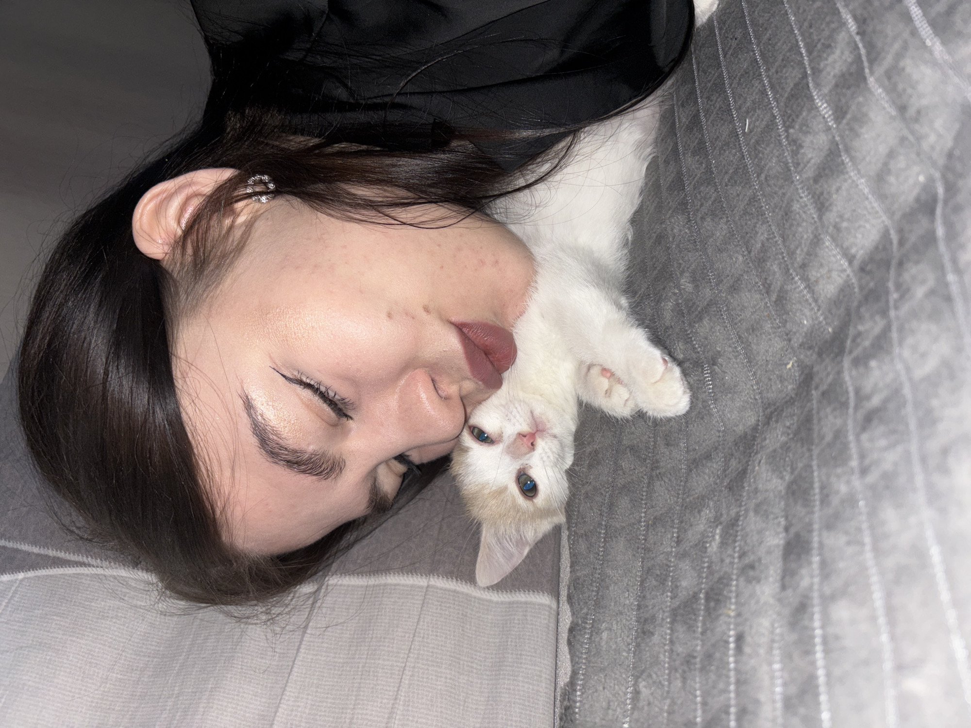 This indoor photograph, taken in Perm, Russia, captures a tender moment between a young woman and a small kitten, both lying down on a soft, grey, textured blanket or bedding. The image is oriented sideways, with the woman's head on the left and the kitten's on the right, nestled close to her face.
The woman has dark hair and is wearing a black garment. Her eyes are closed or looking down, and her lips are slightly parted, gently touching the kitten's head, conveying a sense of affection and relaxation. Visible details include subtle eyeliner and eyeshadow, lip color, light skin with freckles and moles, and a small, sparkling ear cuff on her right ear.
The kitten is small with white fur and light ginger/cream patches, particularly on its head and ears. It has striking blue eyes and is looking directly at the viewer. Its front paws are visible near the woman's face. The lighting is dim, suggesting an evening or nighttime setting, contributing to the intimate and peaceful atmosphere. The grey bedding features a faint striped pattern. No text is visible in the image.