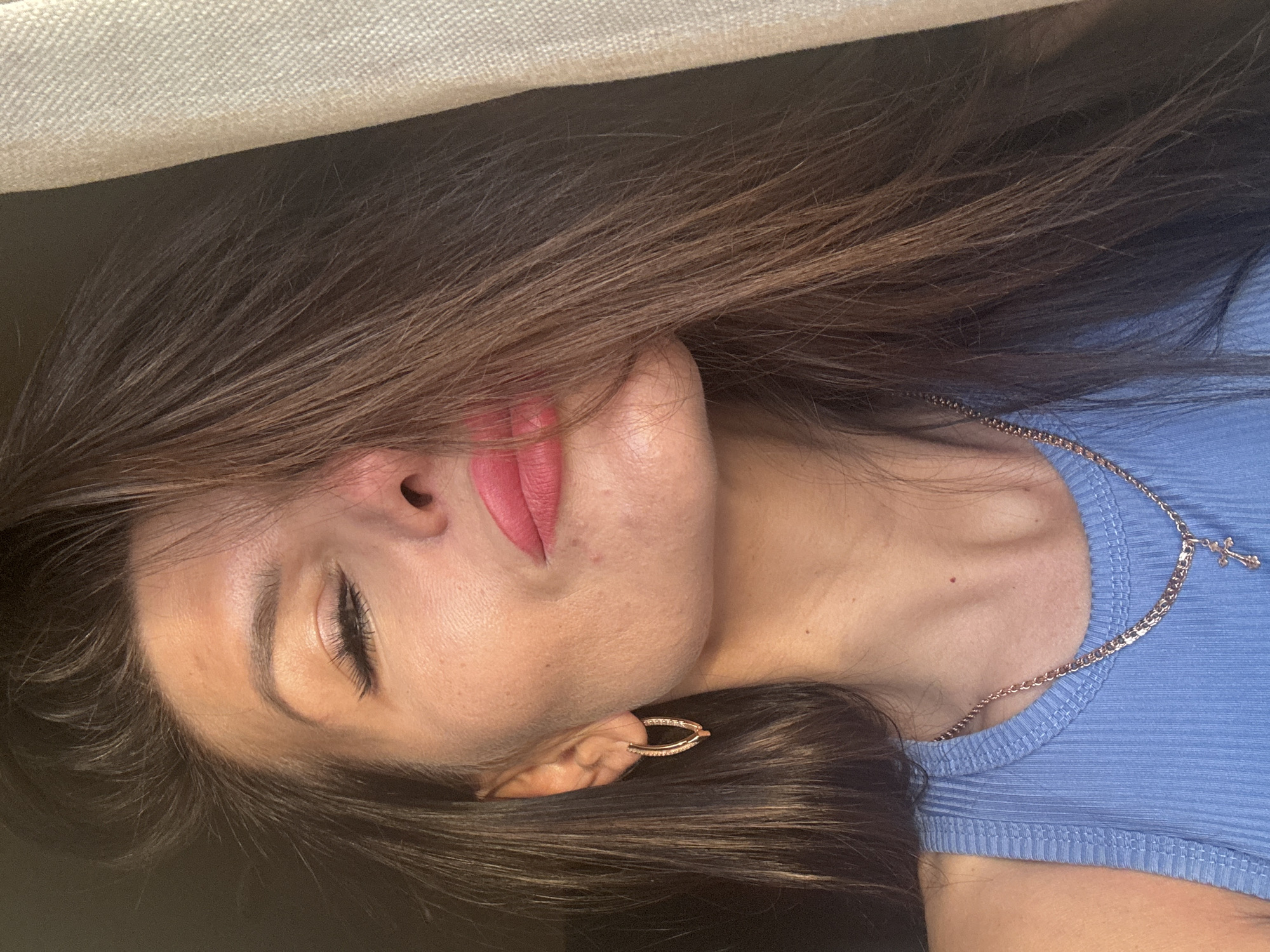 The media file is a close-up, horizontally oriented photograph of a person's face and upper body. The person appears to be lying down, with their head tilted and hair spread out. They have closed eyes, and their lips are colored with a matte pink lipstick. A delicate rose gold chain necklace with a small cross pendant is visible around their neck, resting on a light blue ribbed tank top. A small, sparkling earring is worn in their visible ear. The lighting suggests an indoor setting, possibly with soft, natural light. The overall mood conveyed is one of relaxation or peacefulness. No text is visible in the image.
