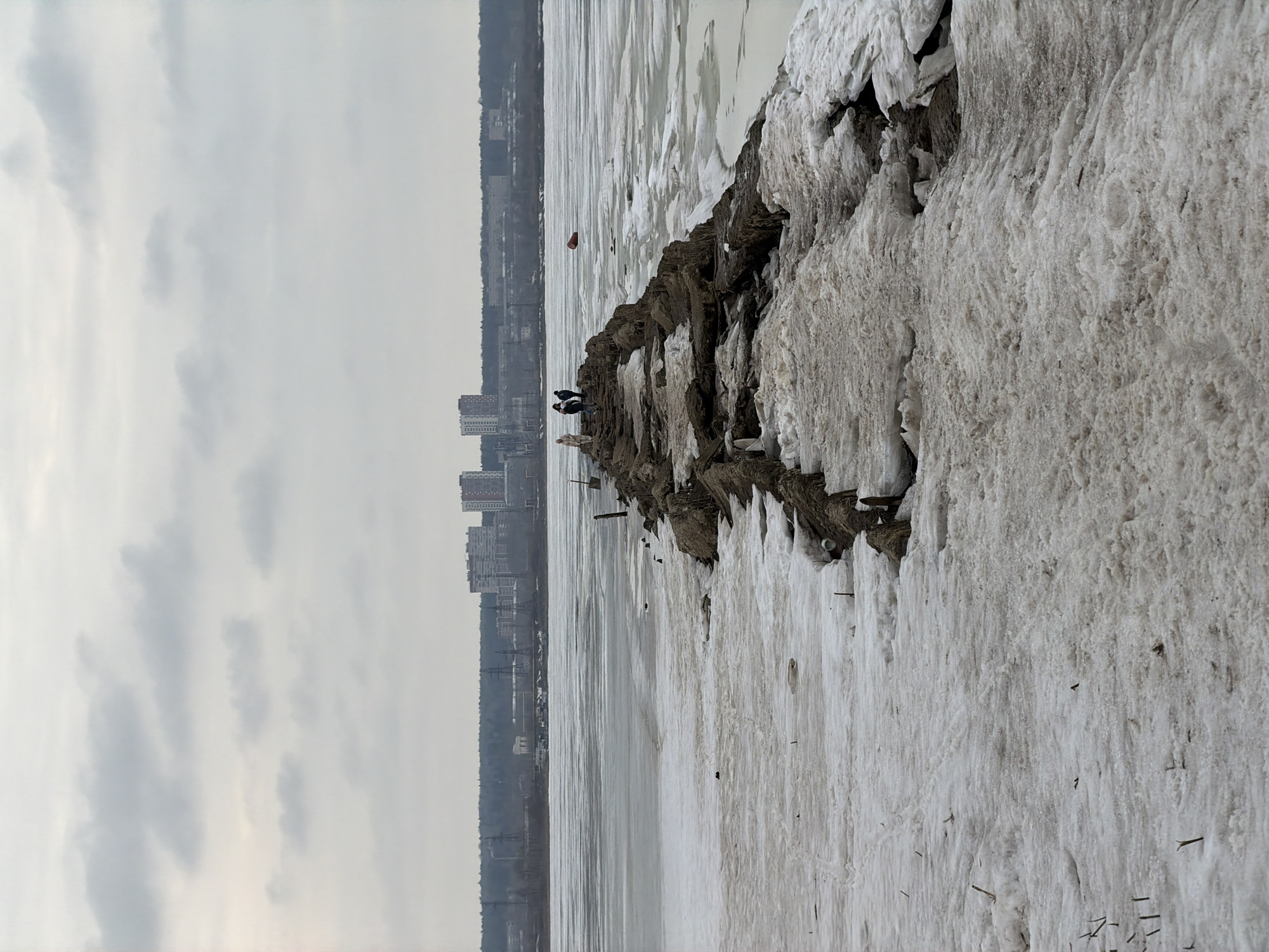 This is an outdoor, slightly high-angle shot of a shoreline in Izhevsk, Russia. The scene appears to be from late winter or early spring, with remnants of snow and ice prevalent. The sky is overcast.
A breakwater or a natural formation of earth and rock runs into the image, partially covered with melting snow and ice. Two people are standing on the structure, visible at a distance. In the background, across the expanse of water, is the skyline of Izhevsk. Several buildings are visible in the city, including residential blocks and industrial structures. The ground leading up to the city seems forested.
In the water, there are patches of open water visible amidst the ice. A red buoy bobs in the distance. Debris like branches and sticks are scattered on the ice and snow.