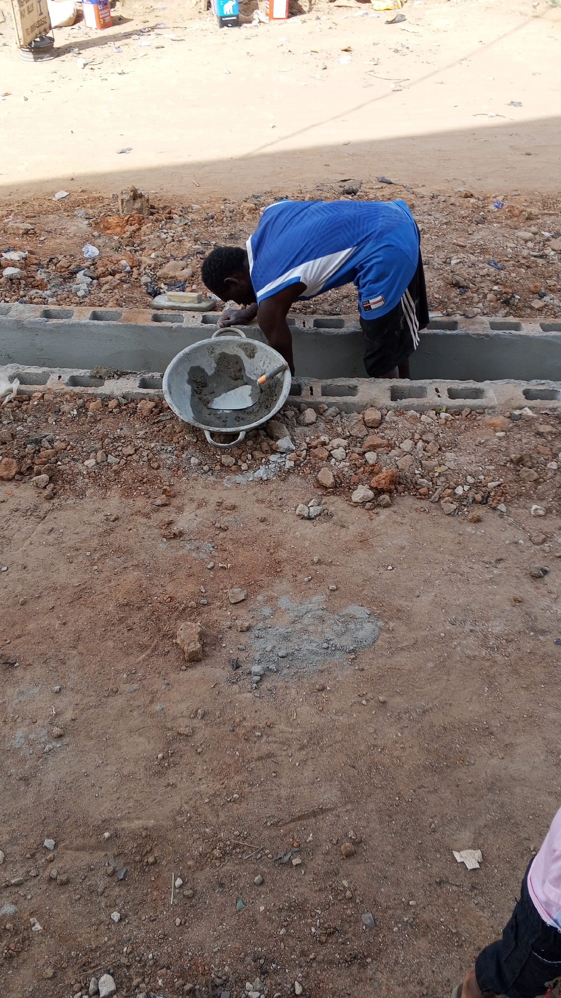 A man wearing a blue and white t-shirt and dark shorts is bending over a grey tub filled with cement and a trowel, appearing to be using the cement to lay a foundation constructed of grey cinder blocks. The location is Sabon Tasha, Nigeria, evidenced by the distinctive red-brown soil and construction style. The ground around the foundation is covered in loose dirt, rocks, and some scattered debris. The background shows a sandy area with some trash and objects in the distance. A partial view of another person's leg and clothing is visible in the bottom right corner.
