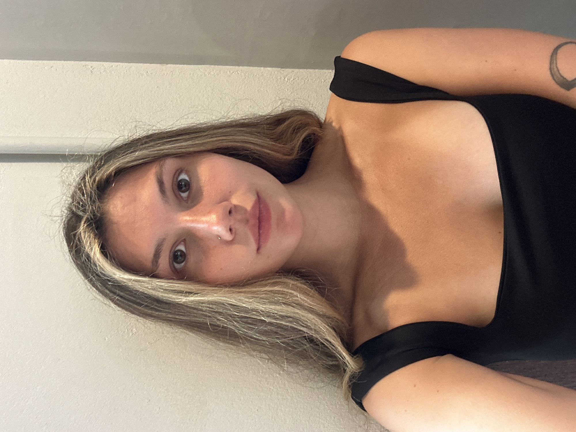 A young woman with fair skin, light brown eyes, and long blonde hair is posing for a selfie. She is wearing a black, off-the-shoulder top. A crescent moon tattoo is visible on her upper left arm. She has a nose piercing. The background is a plain, light-colored wall. The image is oriented sideways. There is no information to indicate the image was taken in Baan Bo Phut, Thailand.
