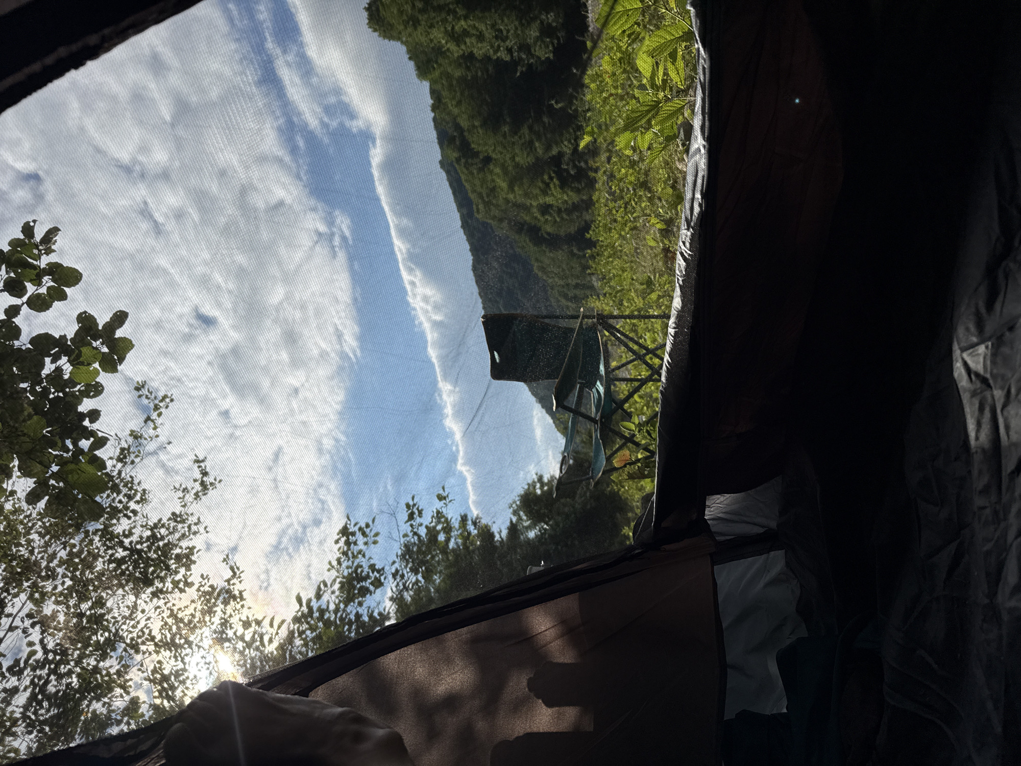 The image shows an outdoor scene viewed from inside a tent. The foreground is dominated by the dark interior of the tent, with fabric textures visible. To the left, a hand is visible, suggesting someone is inside the tent. Sunlight streams through the mesh of the tent opening, illuminating trees with green leaves. Beyond the trees, a steep, lush green mountainside rises under a blue sky with scattered white clouds. A camping chair is visible set up outside the tent, positioned in front of the mountain. The lighting suggests it is daytime, possibly morning or late afternoon due to the angle of the sun. The overall impression is one of a peaceful, natural setting, likely a campsite in a mountainous, vegetated area. The location context provided is Abgara, Abkhazia, which aligns with the mountainous and forested landscape depicted.