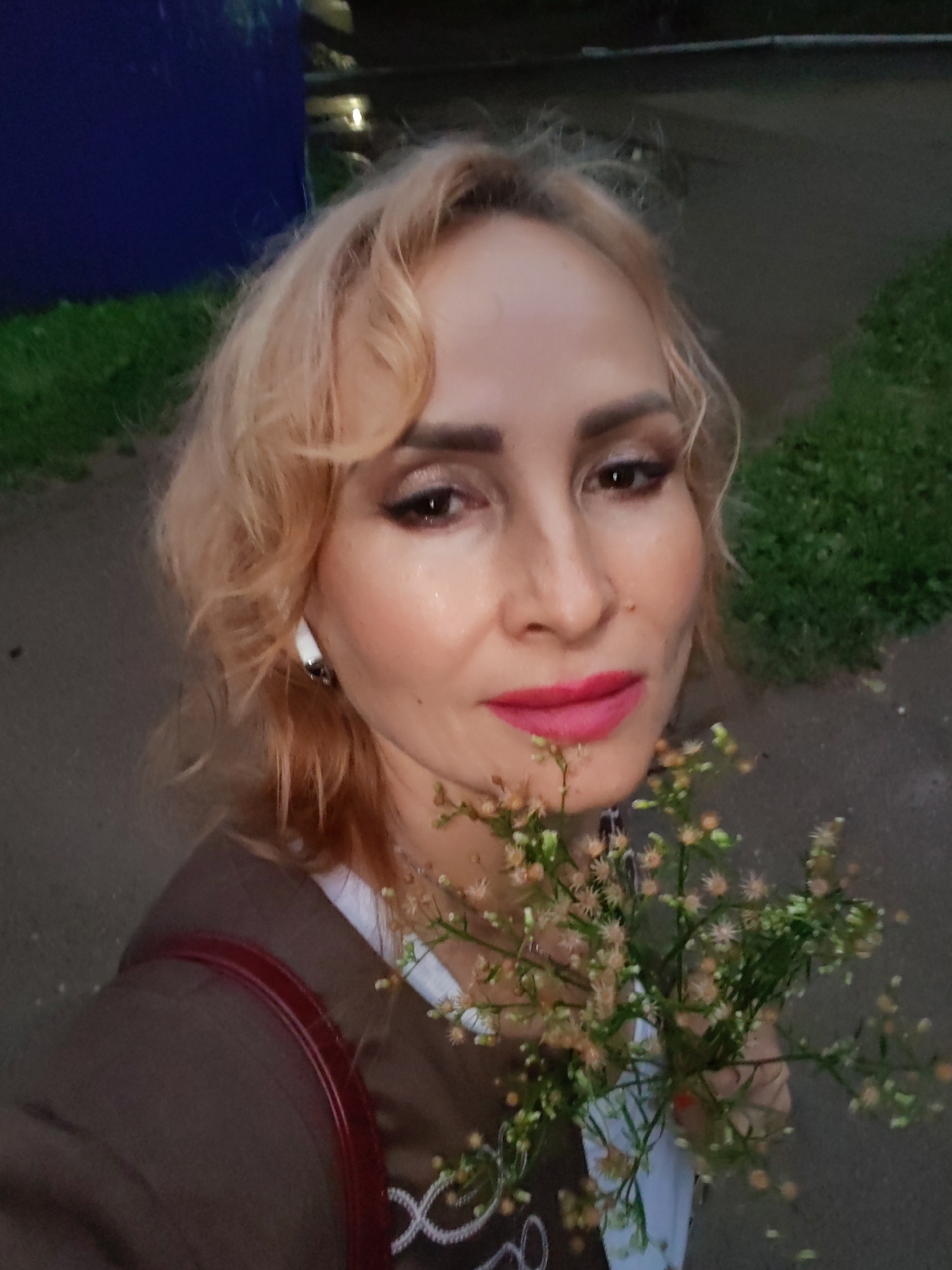 A woman with blonde, wavy hair, fair skin, and bright pink lipstick takes a selfie. She is wearing a brown jacket over a white shirt with some white decorative stitching. She has a red strap over her shoulder, possibly from a purse. She is holding a small bouquet of wildflowers with small, light-colored blossoms and green stems. Her left ear has a white earring. The background appears to be an outdoor setting with a dark asphalt path and green grass on the right. On the left, a large, dark blue structure is visible. The lighting suggests it is dusk or evening. The image was taken in Chaikovsky, Russia.
