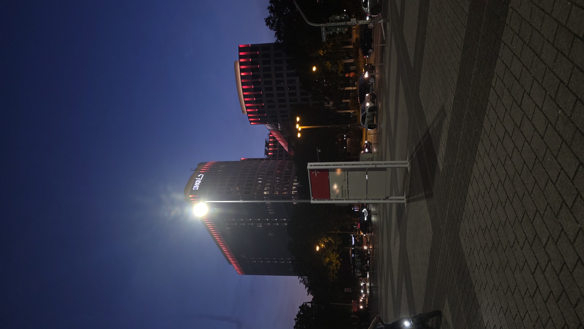 In an urban setting at dusk, two modern office buildings with illuminated red accents on their upper levels dominate the background. The left building features a prominent "ING" logo. In the foreground, a streetlamp shines brightly to the left of the taller building. A multi-panel sign with text and an arrow is positioned centrally. The street is wet and reflects the lights, with several cars and a bus visible, suggesting moderate traffic. The ground is paved with interlocking bricks. The sky is a deep blue, transitioning from twilight hues.