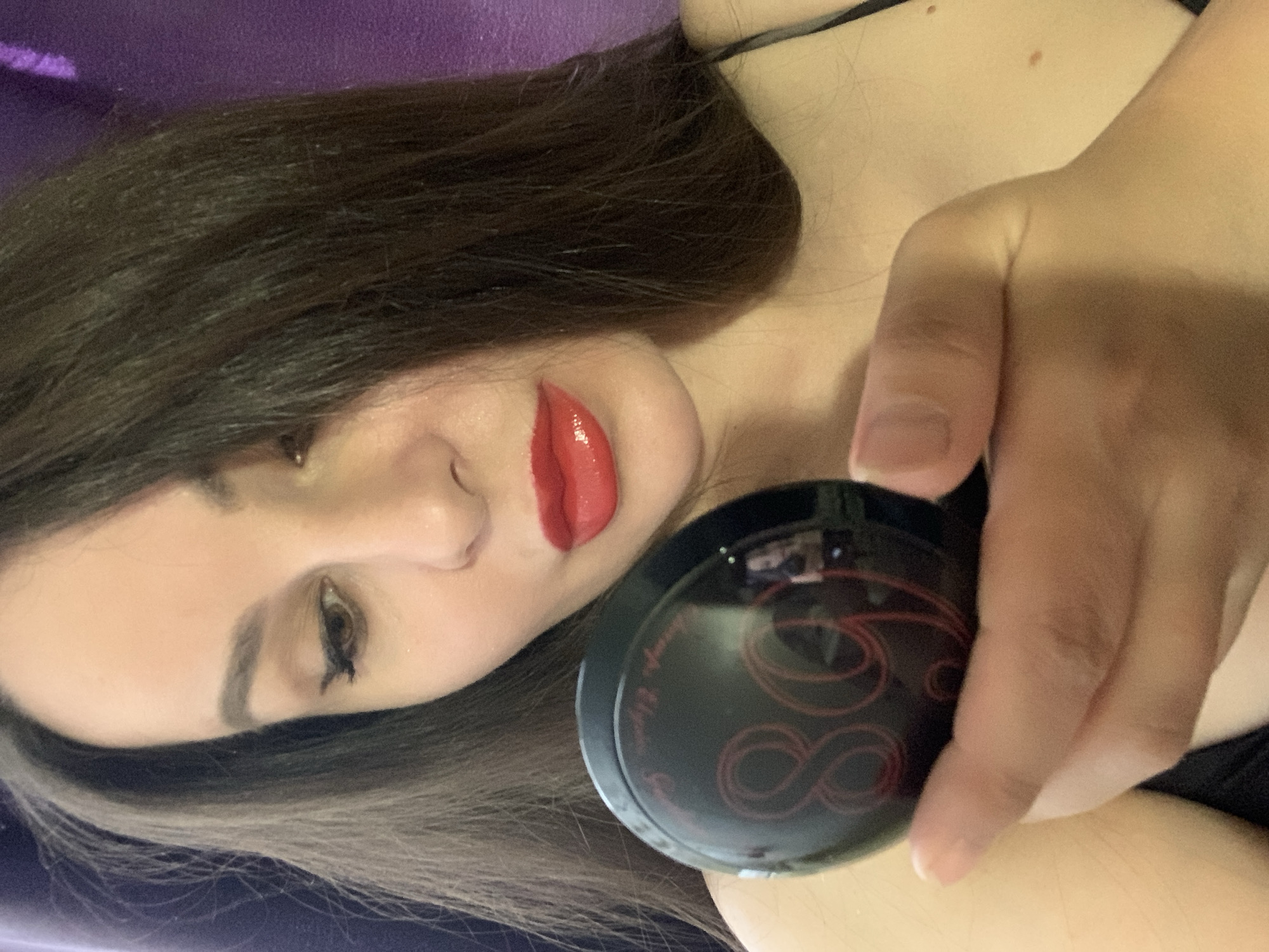 A person, appearing to be a woman with long, dark brown hair, is depicted in a close-up, rotated image. She is lying down, with her head tilted to the side, looking towards the camera with a neutral expression. Her right eye is visible, featuring dark eyeliner and mascara, and her eyebrows are well-defined. Her lips are coated with a prominent, glossy red lipstick. She is wearing a dark-colored top, indicated by a thin black strap visible near her neck and shoulder.
In her right hand, she holds a round, black compact or container. The compact has a reflective surface and is adorned with a red, stylized infinity symbol (or potentially the number "88") and some text. The visible text on the compact appears to read "L'ange Magic" in a cursive font. The background consists of a solid purple fabric, suggesting she is reclining on a bed or a soft surface. The lighting appears soft and even, highlighting her features and the product she is holding.