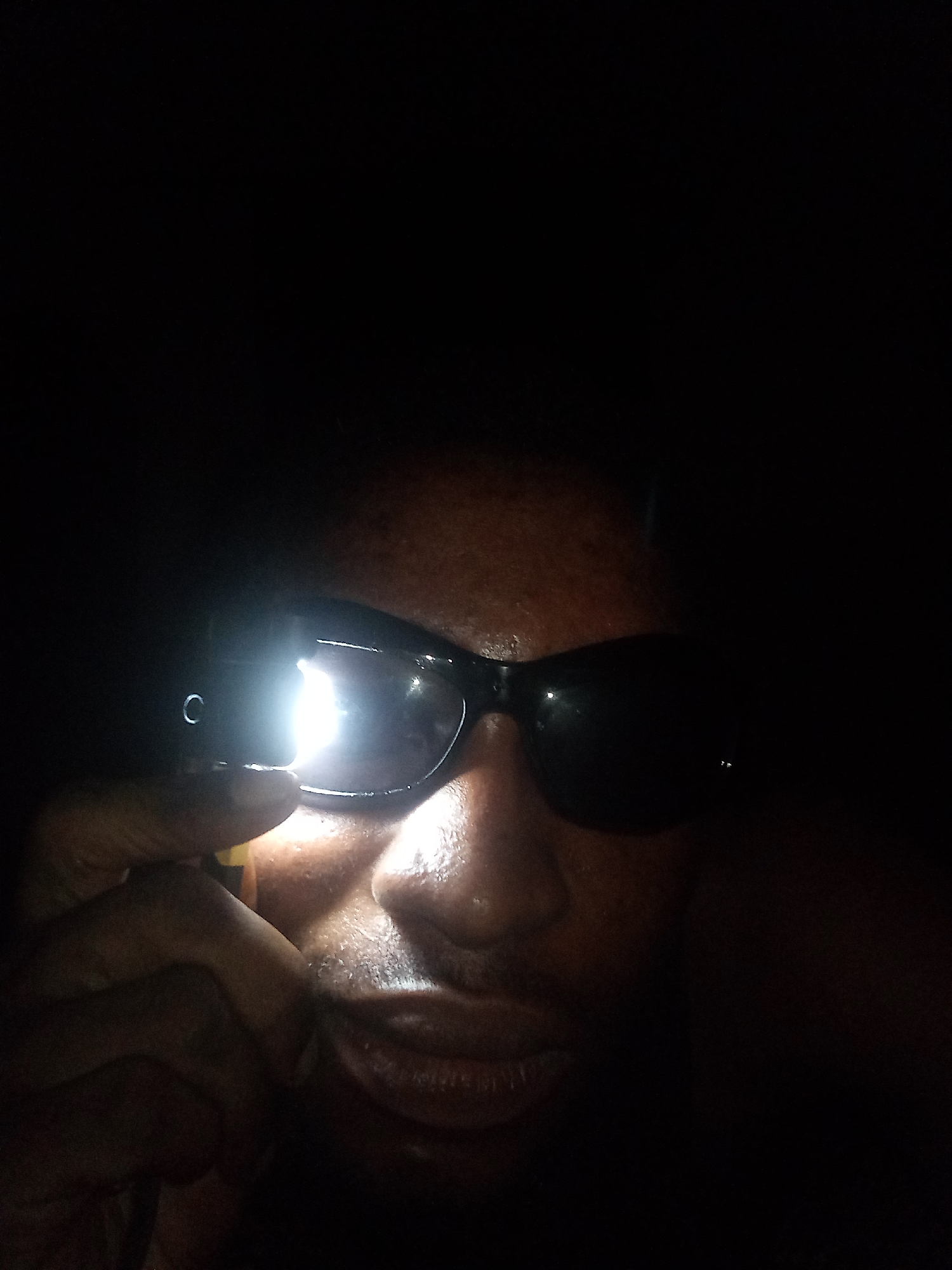The image shows a person with dark skin wearing black sunglasses. A light source, possibly from a phone or small flashlight, is directed at the sunglasses, creating a bright reflection on one of the lenses and illuminating part of the person's face. The background is dark. It is not possible to verify the location of Lokoja, Nigeria from the image.