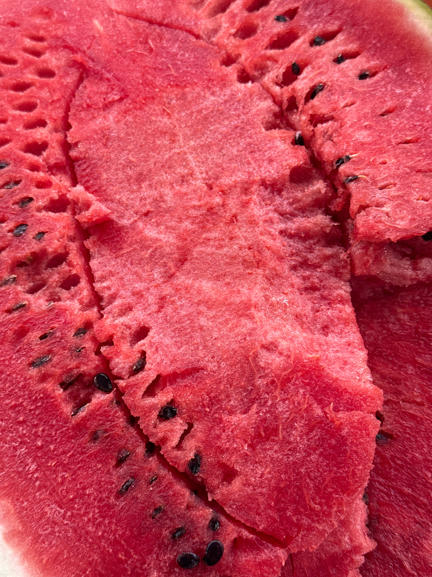 A close-up, top-down view of a sliced watermelon reveals its vibrant red flesh dotted with black seeds. The texture is slightly grainy, with some areas appearing more fibrous and others smoother. Some of the seeds are partially submerged in the flesh, while others are more exposed. The lighting creates subtle shadows and highlights, emphasizing the uneven surface of the fruit. No specific location in Lyubertsy, Russia, can be determined from this image.