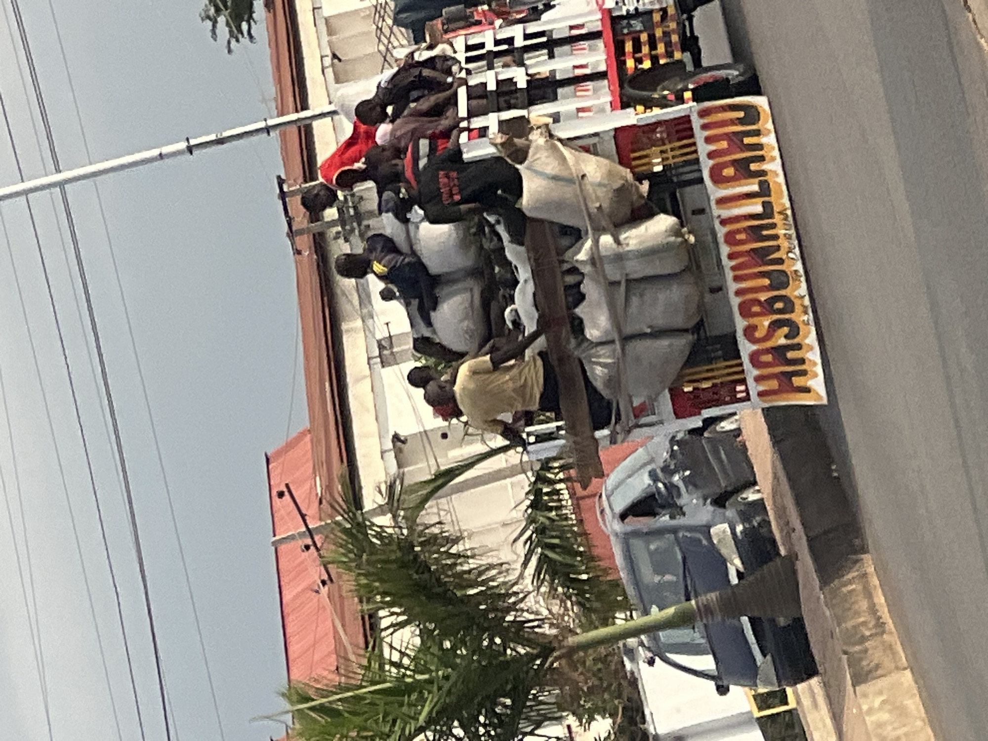 The image shows a vehicle in Barakin Lerawi, Nigeria, overloaded with people and large bundles of material. The sign on the vehicle says "HASBUNALLAHU." The vehicle is on a paved street next to a curb and other parked vehicles, in an urban environment with buildings and a palm tree visible.