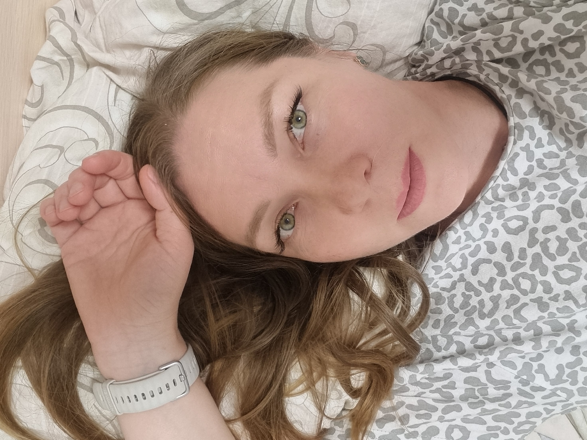 A woman is lying on a bed or similar surface. She is wearing a grey and white animal-print shirt. She has brown hair and green eyes. Her left hand is raised and resting near her head, and a white watch band is visible on her left wrist. She appears to be in an indoor setting with patterned bedding. The location in the city of Chaikovsky, Russia is not discernible from the image.