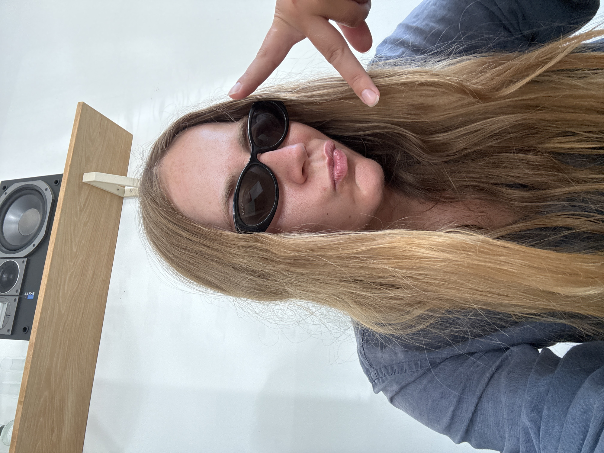 A young Caucasian woman with long, blonde hair is pictured in a selfie. She is wearing black, oval-shaped sunglasses and a blue-gray collared shirt. Her lips are pursed, and her right hand is raised, with her index and middle fingers extended in a peace sign. The background is a plain white wall with a wooden shelf mounted above her. On the shelf, a black speaker is visible. The scene appears to be indoors, likely in an apartment or studio, and there is no indication of a specific location in Đà Nẵng, Vietnam.