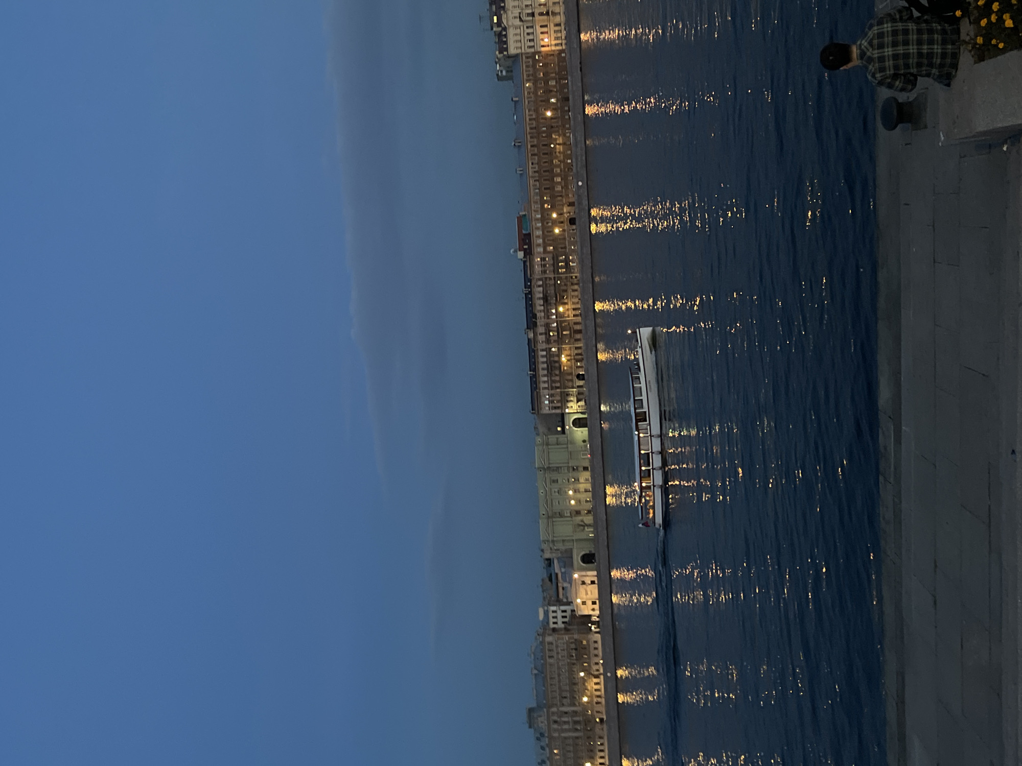 The image depicts a dusk scene along the Neva River in Saint Petersburg, Russia. On the far bank, a row of classical buildings, illuminated by numerous lights, stretches across the horizon. A white passenger boat with its lights on navigates the dark water. The river's surface reflects the lights from the buildings and the boat, creating shimmering trails. In the foreground, a person wearing a plaid shirt is seated on a stone embankment, facing the river and the distant cityscape. The sky above is a deep, clear blue. The overall atmosphere is serene and reflective.