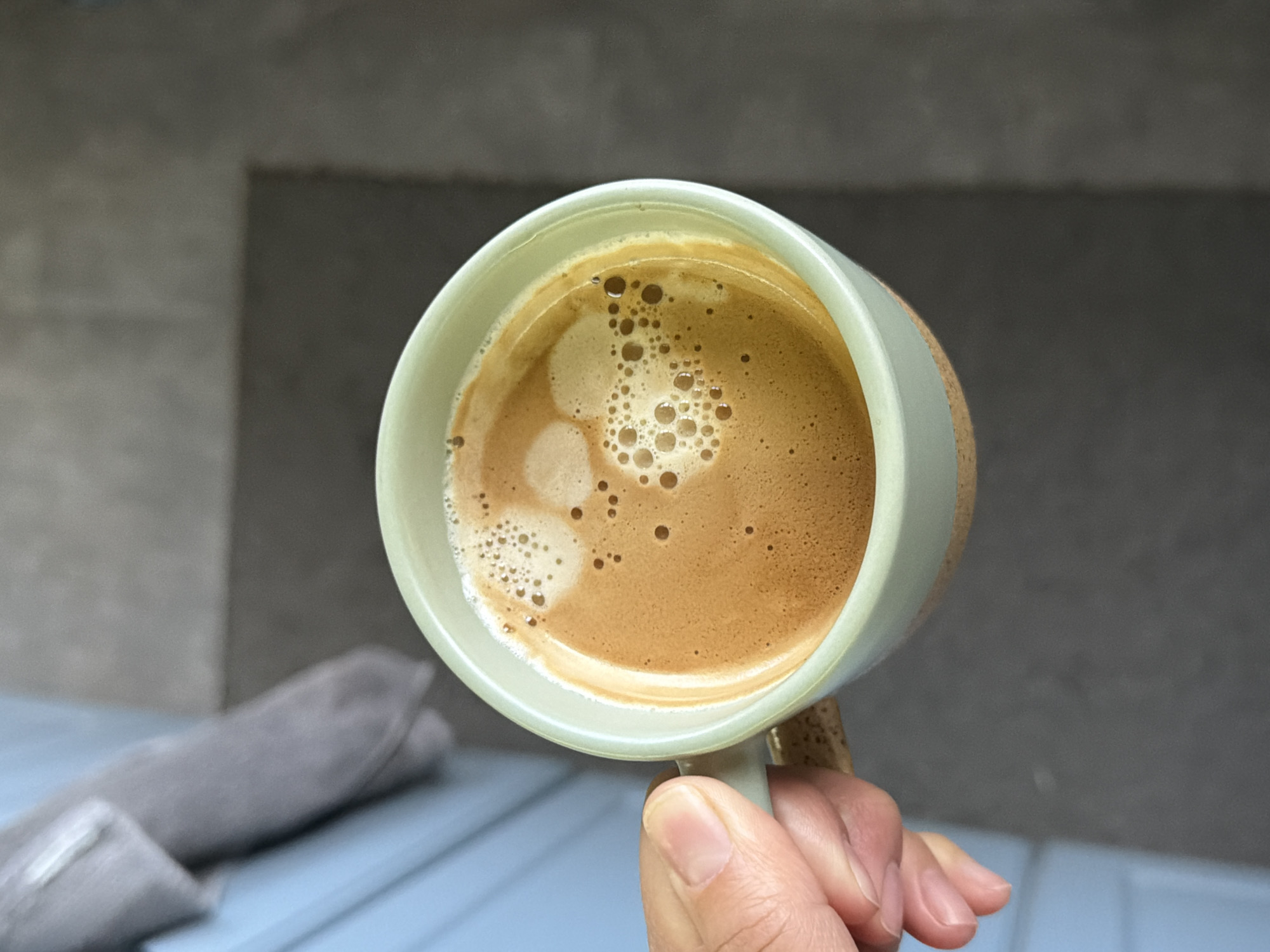 A close-up, eye-level shot shows a hand holding a mug filled with a coffee-like beverage. The mug is light-colored with a light green rim. The beverage has a tan color with small and large bubbles on the surface. The hand holding the mug is light-skinned. The background consists of a gray-painted wall and a blue surface with a gray cloth object resting on it. There is no information to suggest this image was taken in Pleven, Bulgaria.