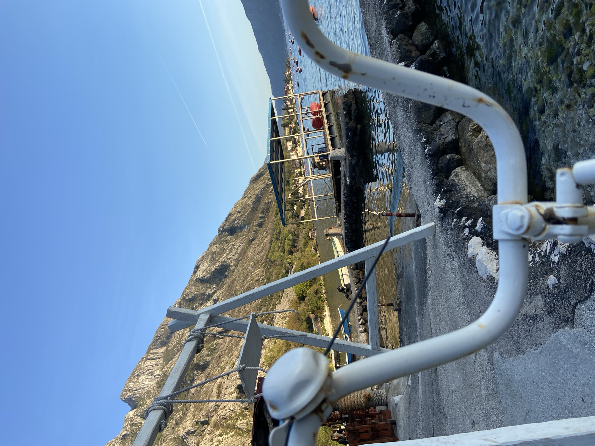 The image shows a view from a dock in Donji Orahovac, Montenegro, looking out towards the Bay of Kotor. In the foreground are sections of white metal railings and structural supports of the dock. The bay is visible, with a small floating platform marked by red buoys. Across the bay, a mountain rises sharply from the water's edge, and a settlement with buildings is visible along the shoreline. The sky is clear and blue, with a contrail from an aircraft visible. The water along the dock's edge is clear, with visible rocks and pebbles. The dock itself appears to be made of concrete.