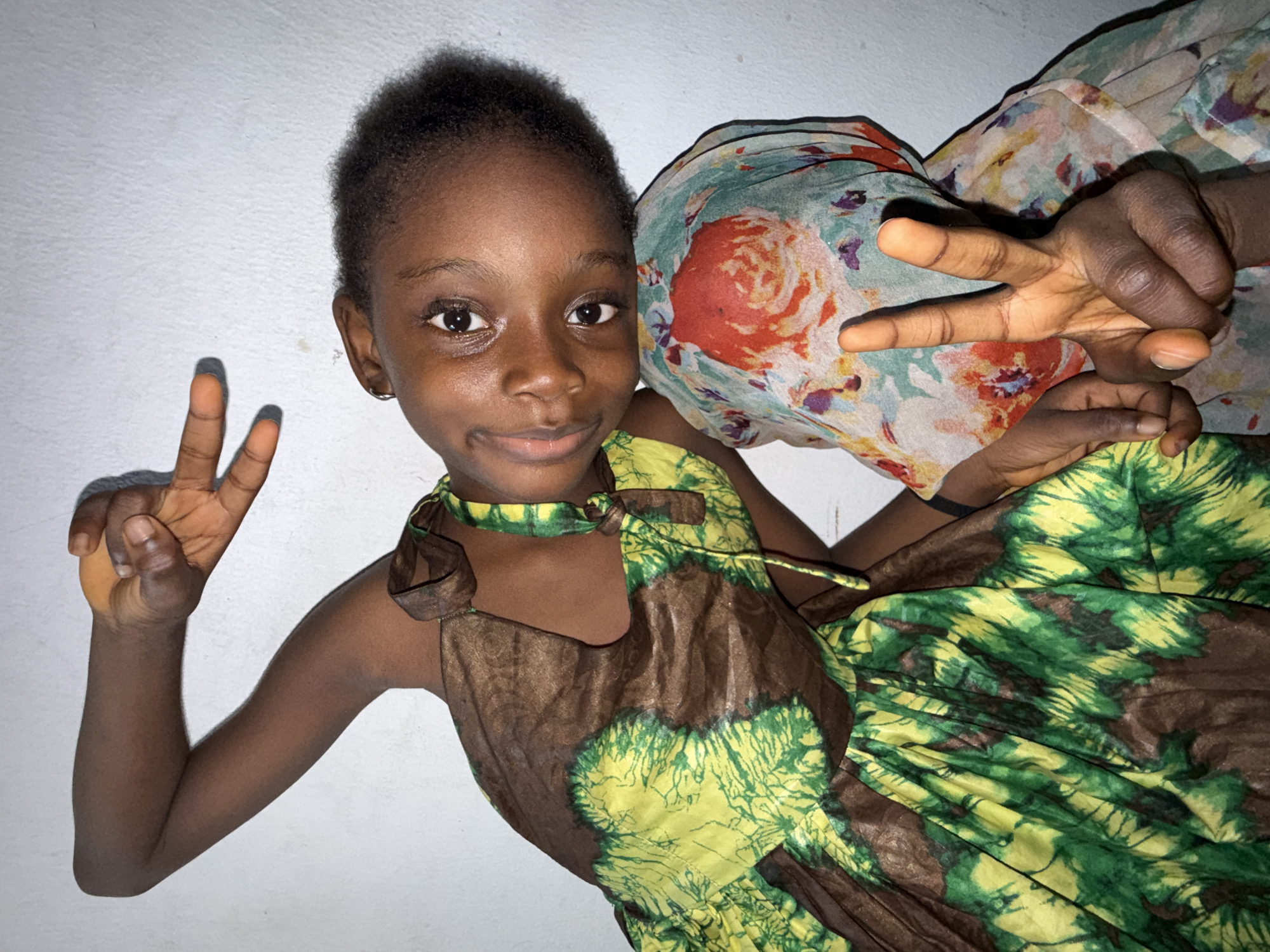 In Ado, Nigeria, a young girl with dark skin is pictured indoors, leaning against a light-colored wall. She wears a brown and green tie-dye dress. Her left arm is raised, with her hand forming a peace sign. Her head is tilted, and she is looking towards the camera with a slight smile. Another person, or a part of another person, is partially visible to her right, wearing a headscarf with a floral pattern. This second individual's hand is also visible, forming a peace sign with their fingers. The lighting suggests an indoor setting, possibly with artificial light. The overall scene is casual and playful.
