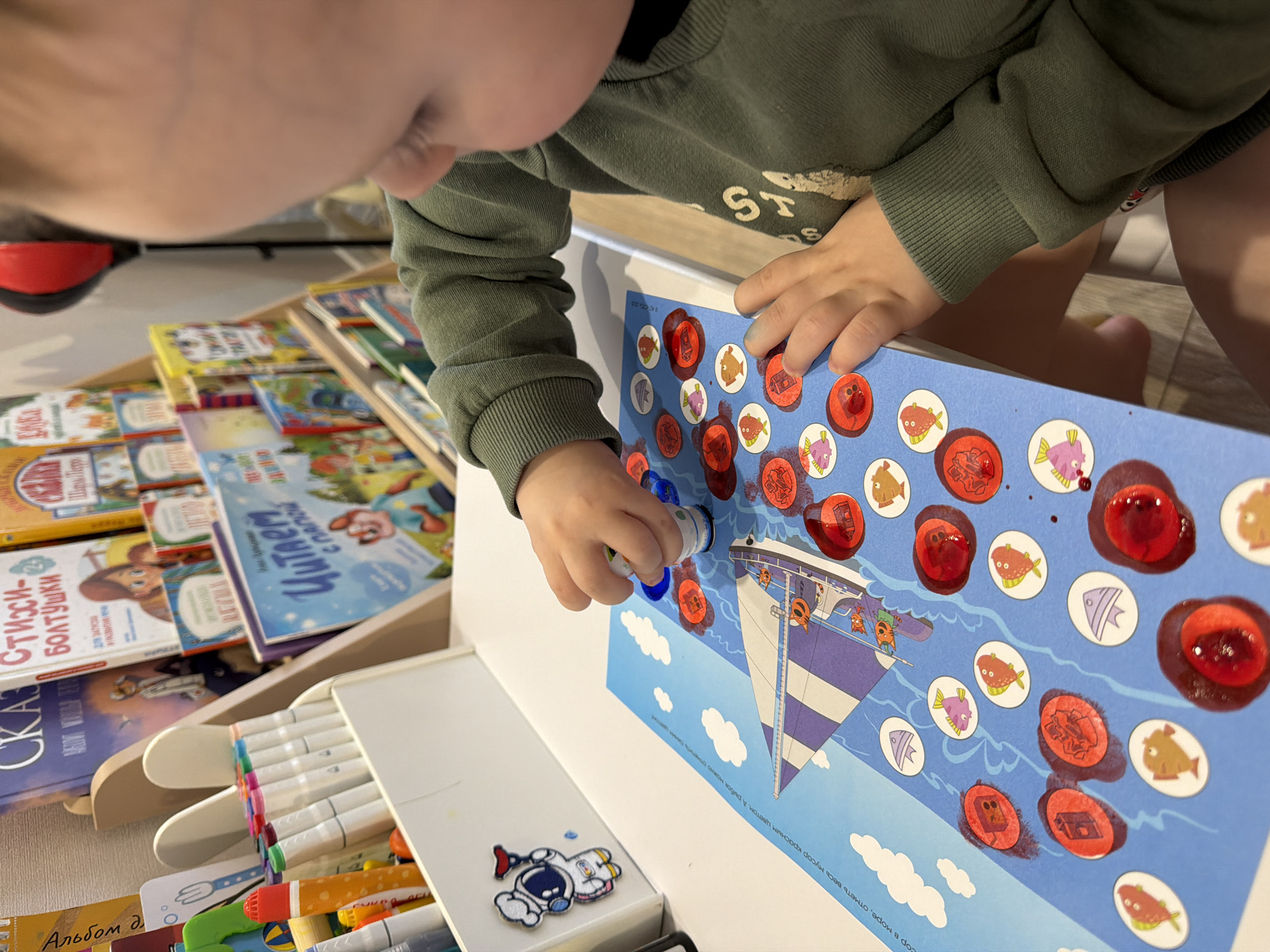 A toddler wearing a long-sleeved green shirt stamps red ink onto a blue educational poster depicting a boat sailing on water and surrounded by circles containing fish. The activity appears to be related to marking trash in the sea with red ink. The toddler's hands and face are visible. A collection of books and markers sits to the left of the poster. The location is likely indoors in a private residence, potentially in Chaikovsky, Russia.