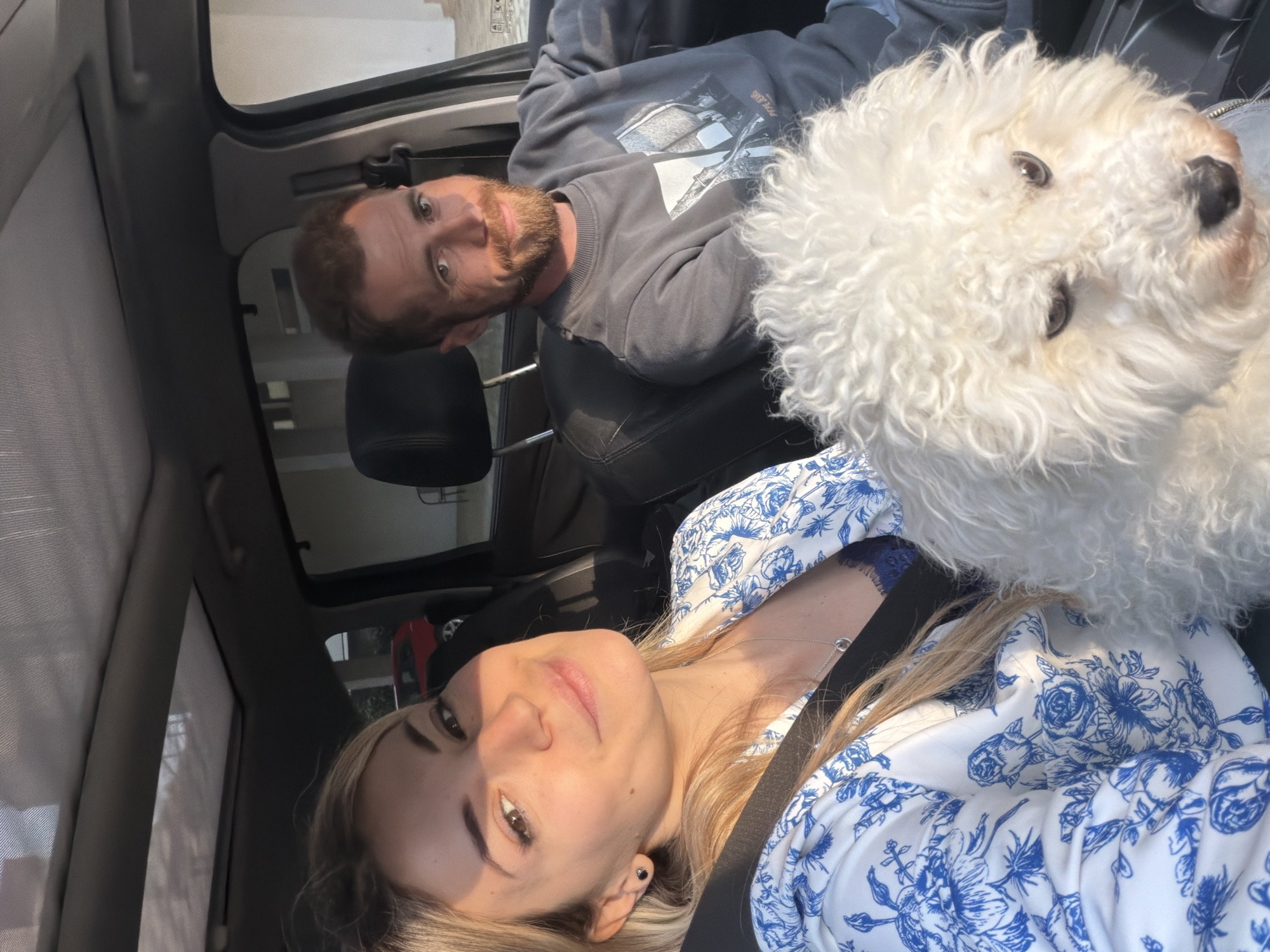 The image is a sideways-oriented selfie taken inside a car, featuring two people and a white fluffy dog, captured during a bright, sunny period, likely late afternoon or early evening as suggested by the warm, directional lighting. The setting is a car interior, with grey seats and headrests visible.
On the left, a man with a beard and short brown hair is seated, wearing a dark grey long-sleeved sweatshirt with a graphic print and the brand "PEPE JEANS" visible. He looks directly at the camera with a neutral expression. Next to him, a white, curly-haired dog, possibly a Bichon Frise or Poodle, is featured prominently, looking alertly towards the camera. On the right, a woman with long blonde hair and a pleasant, slight smile is visible. She is wearing a white blouse adorned with a distinctive blue floral pattern and a silver necklace, with a car seatbelt crossing her chest.
The group appears to be posing for a casual photo, possibly during a trip or outing in Budva, Montenegro. The atmosphere is relaxed, with the subjects appearing comfortable in their environment.
